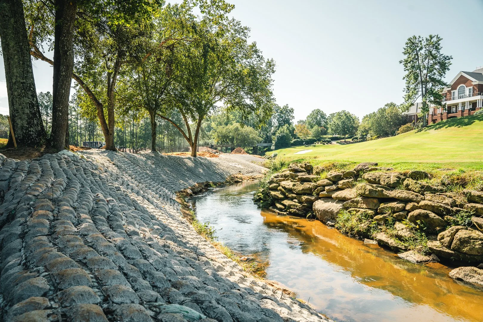 A peaceful scene of a small stream running alongside a stone embankment, with trees providing shade and a lawn with a large house in the background.