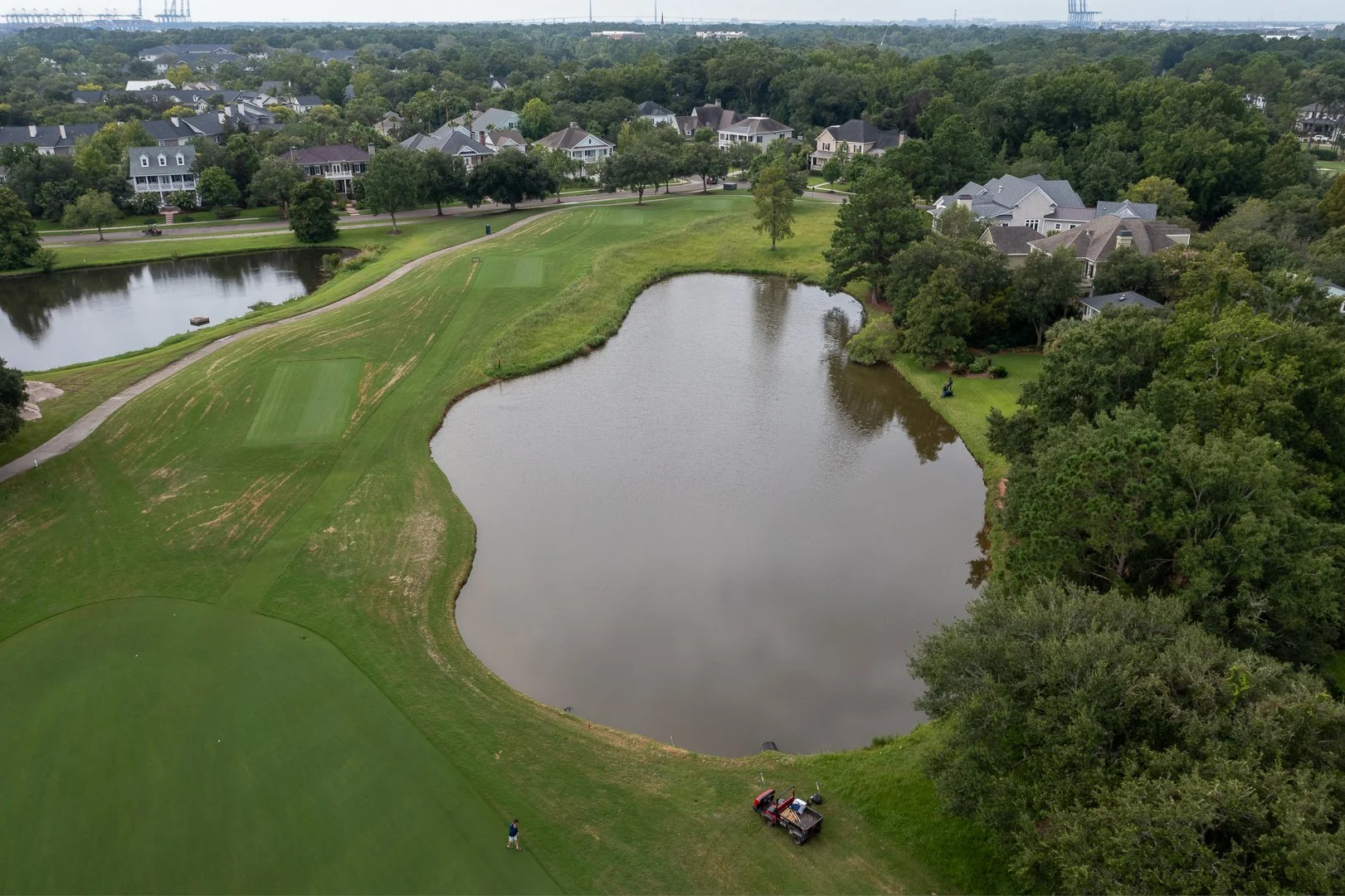 Aerial view of a golf course with a pond, surrounded by trees and residential houses in the background.