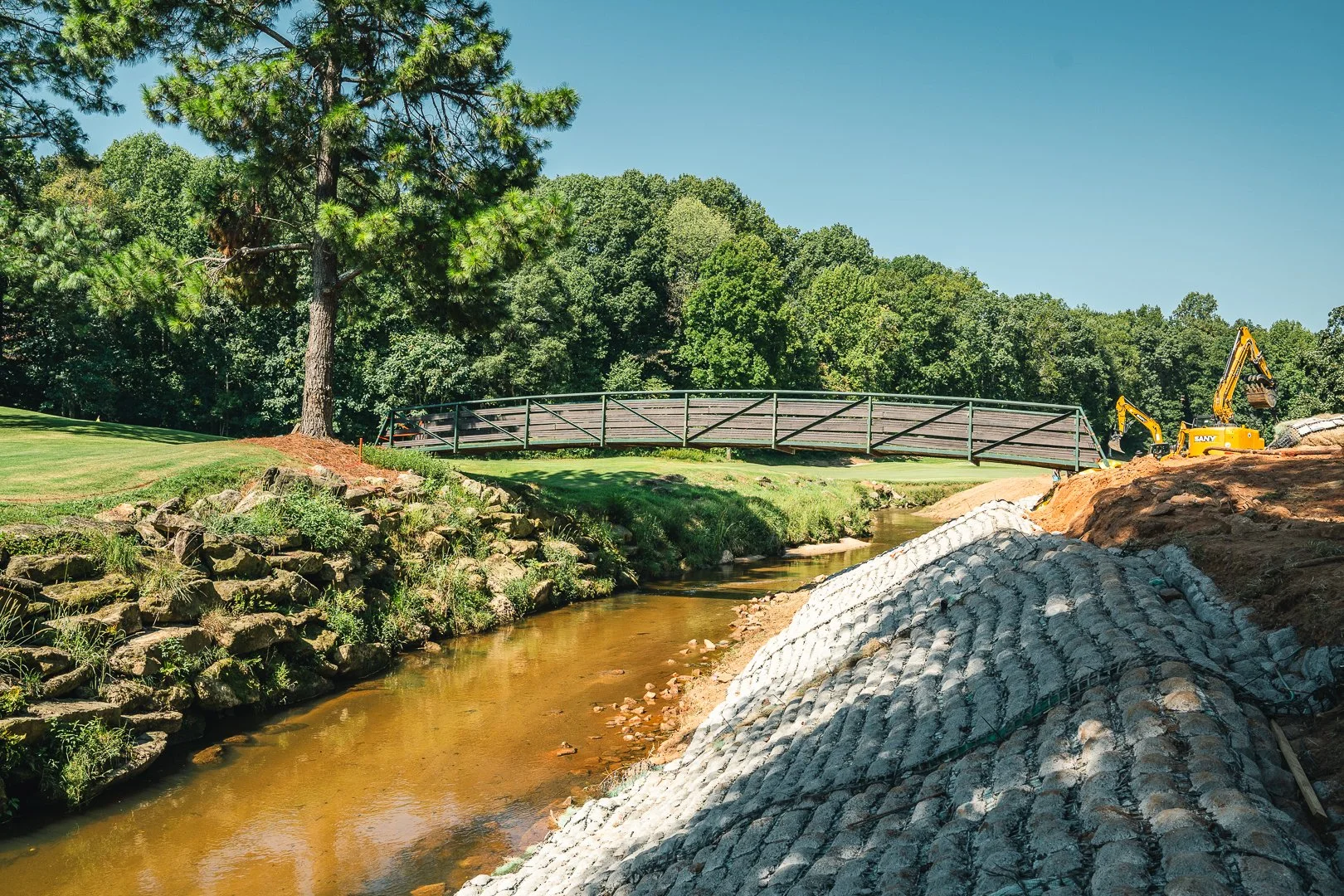 Construction site near a small creek with a bridge, trees, and machinery.