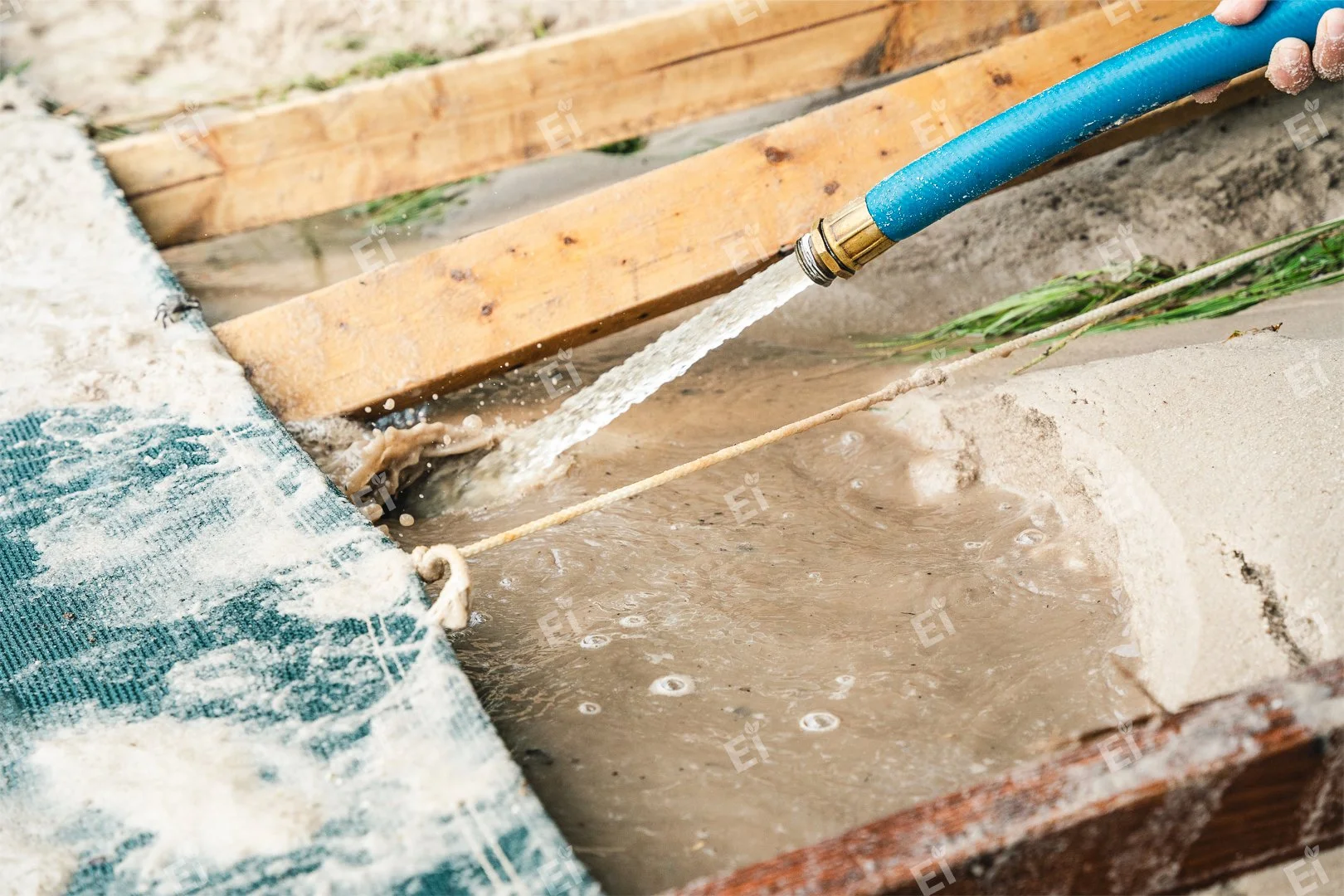 A blue garden hose is spraying water into a concrete area surrounded by wooden frames and sand, with a piece of string stretched across the area.