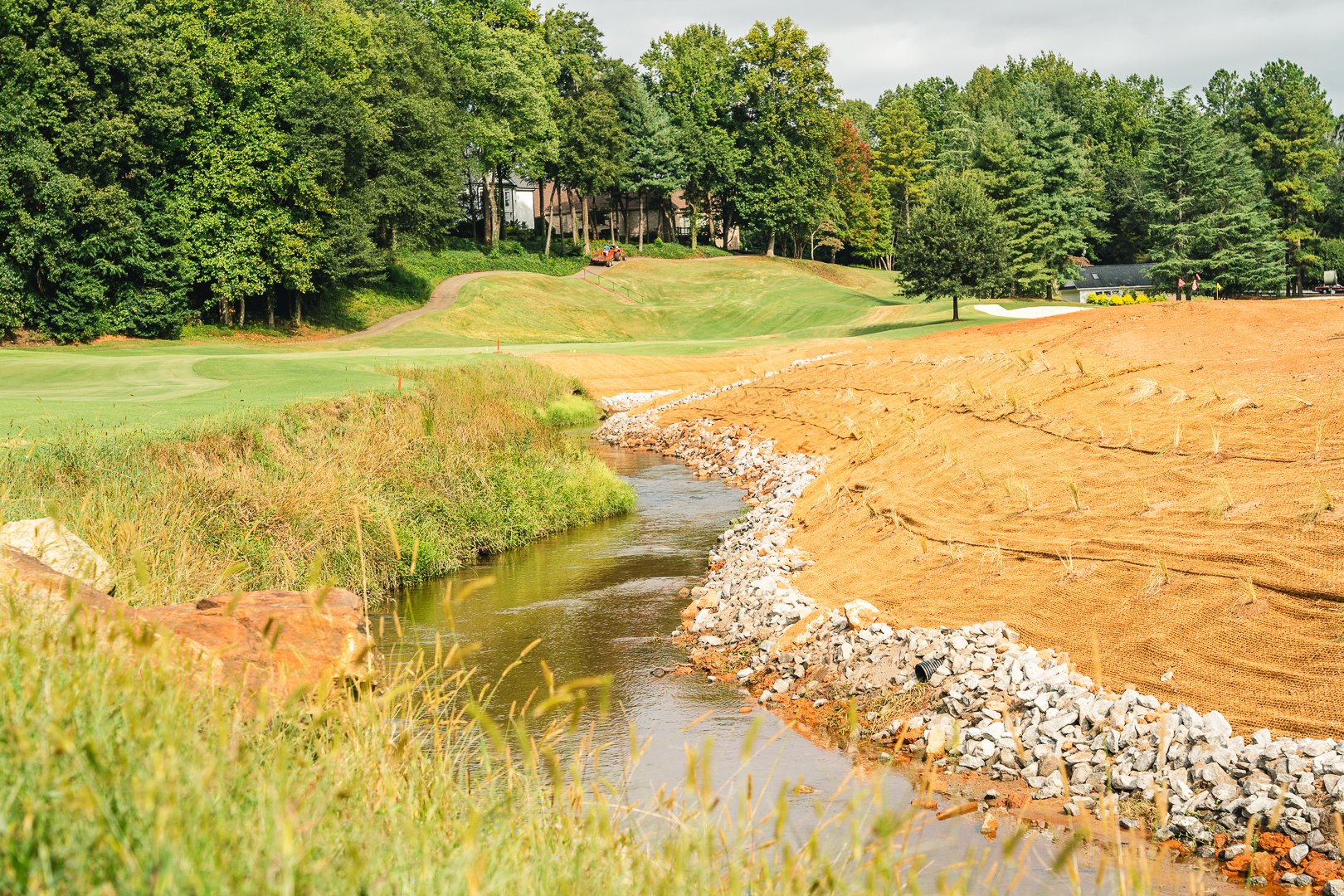 A golf course under construction with a small creek running along the edge, a green fairway in the distance, and trees surrounding the area.