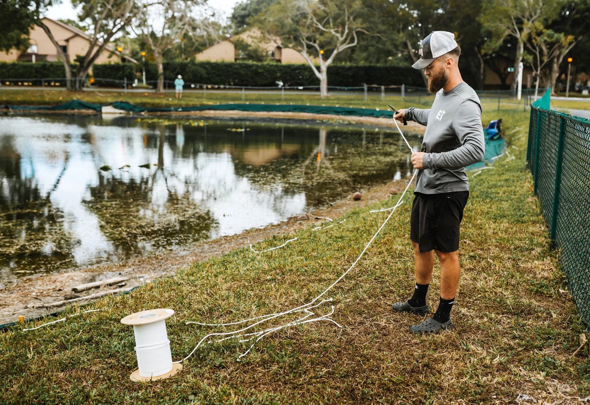 A man wearing a gray long-sleeve shirt, black shorts, and a gray and black cap stands by a pond. He is holding a rope connected to a spool of white string, which is partially unraveled on the ground. The pond has some floating debris and trees reflected in the water. In the background, there are houses, trees, and a person walking on a path.