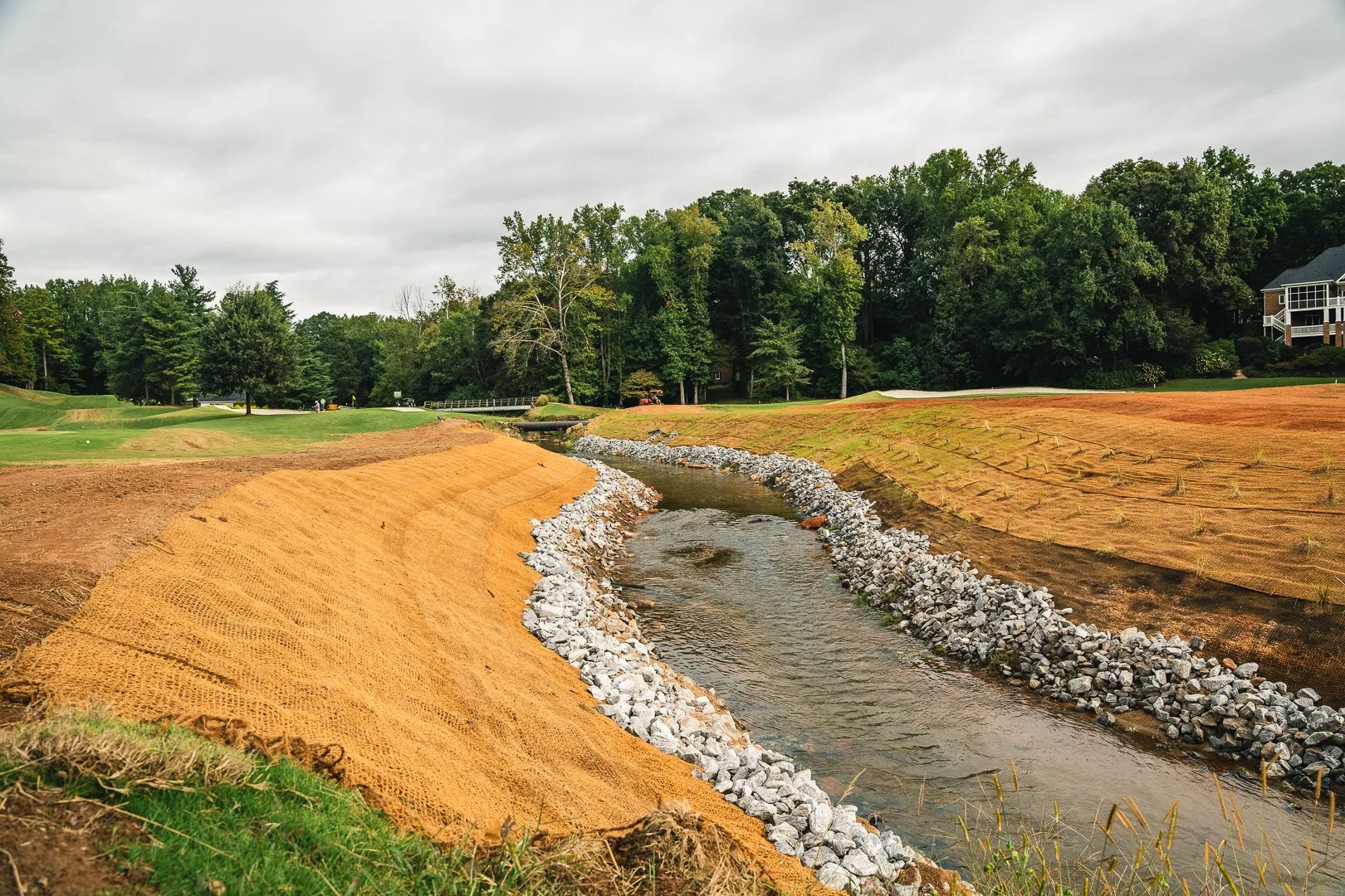 Construction area with a small stream of water, rocks, and orange plastic covering soil, with grassy areas and trees in the background.