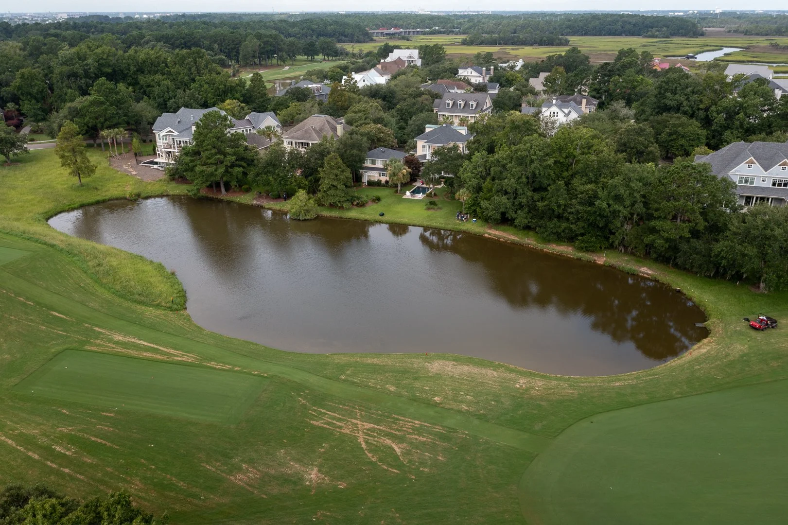 Aerial view of a residential neighborhood with large houses, trees, and a pond or small lake in the center, surrounded by green lawns and fields.
