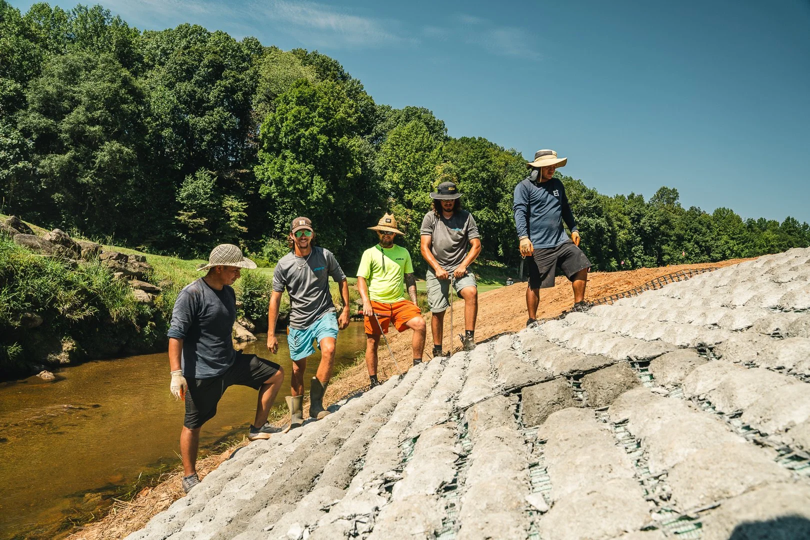 Group of five men wearing hats and casual outdoor clothing walking along a sloped, rocky embankment next to a small stream, surrounded by green trees and blue sky.