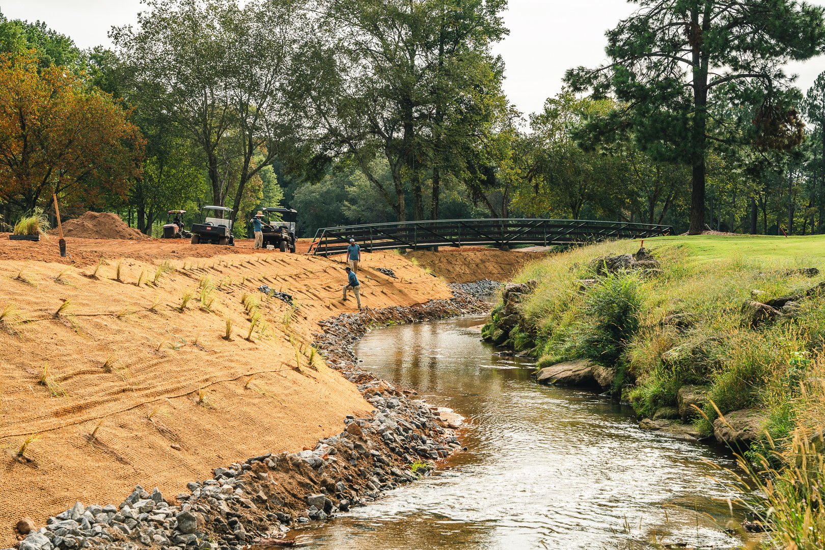 Workers and golf carts on a course near a small stream with a bridge and trees in the background