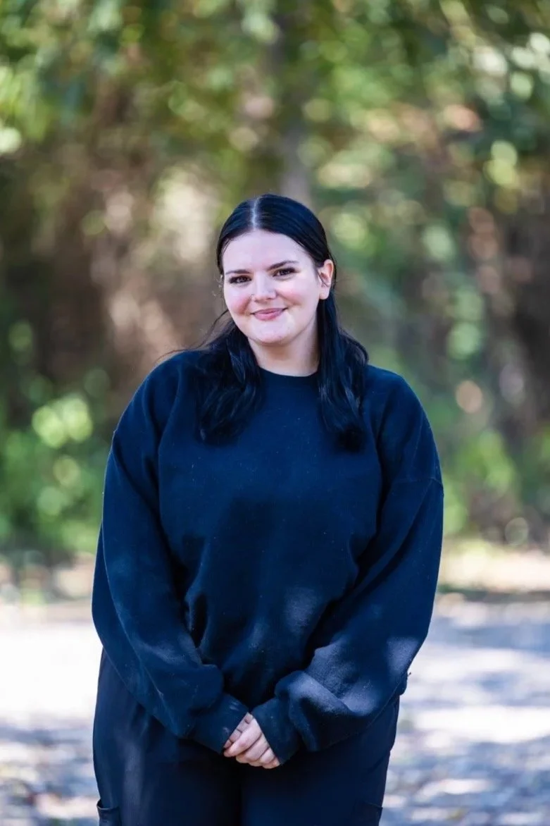 A young woman with dark hair and fair skin standing outdoors in a natural setting, smiling and wearing a black sweatshirt.