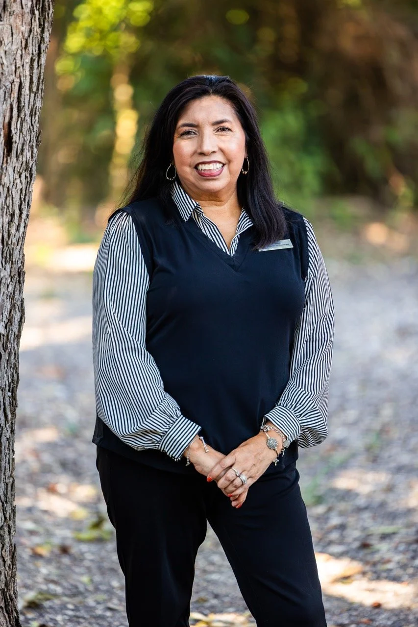 A smiling woman in business attire standing outdoors with trees in the background.