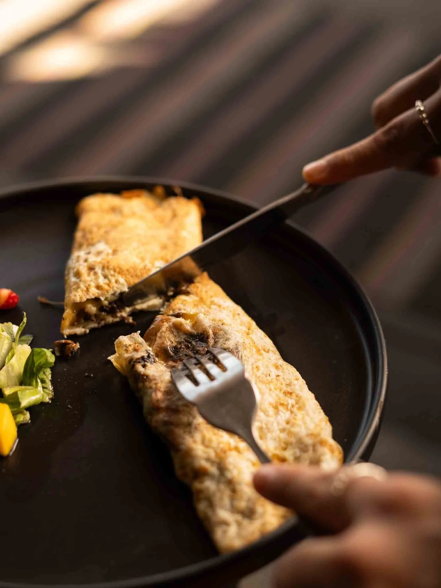 Persona cortando un filete de pescado empanizado en un plato negro, acompañado de ensalada de lechuga, pimientos y tomates. hotel baxar pie de la cuesta 