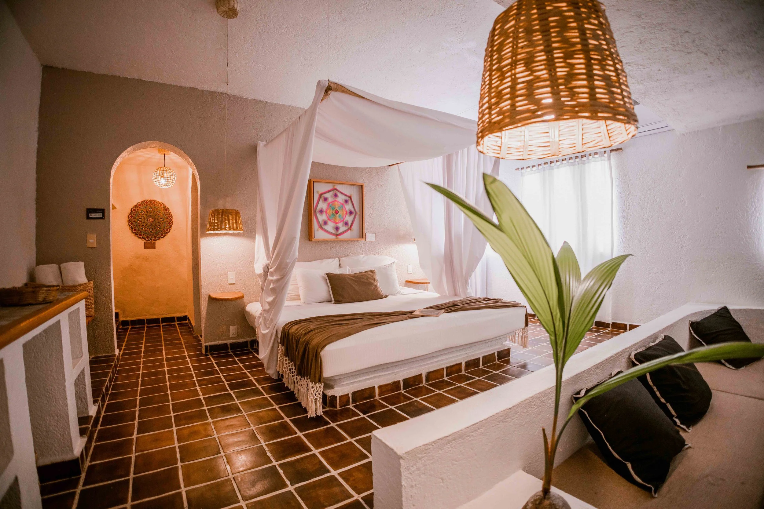 Habitación decorada con estilo rústico, cama de dosel con cortinas blancas, almohadas blancas y una almohada marrón, piso de baldosas marrones, paredes blancas con textura, plantas verdes, lámparas de mimbre, cuadros decorativos en las paredes, y cor