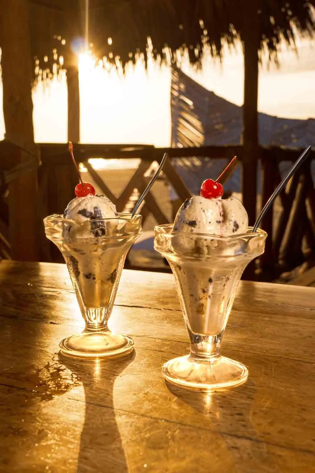 Dos conos de helado de vainilla con trozos de chocolate y una cereza en la parte superior, en copas de cristal en una mesa de madera, con un fondo de una puesta de sol en una estructura de madera con techo de paja.