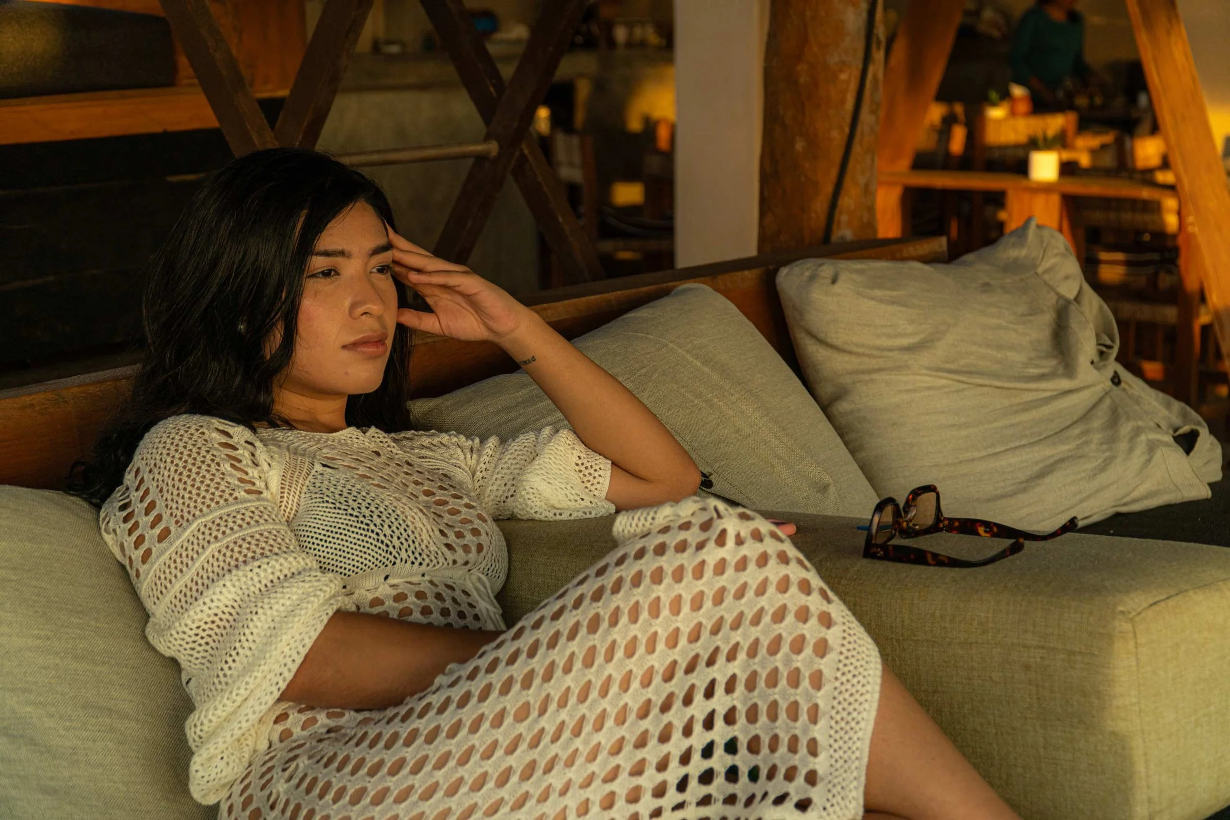 Mujer sentada en un sofá con la mano en la cabeza en un ambiente acogedor, con luz cálida y muebles de madera en el fondo. Hotel boutique playa pie de la cuesta, baXar Acapulco