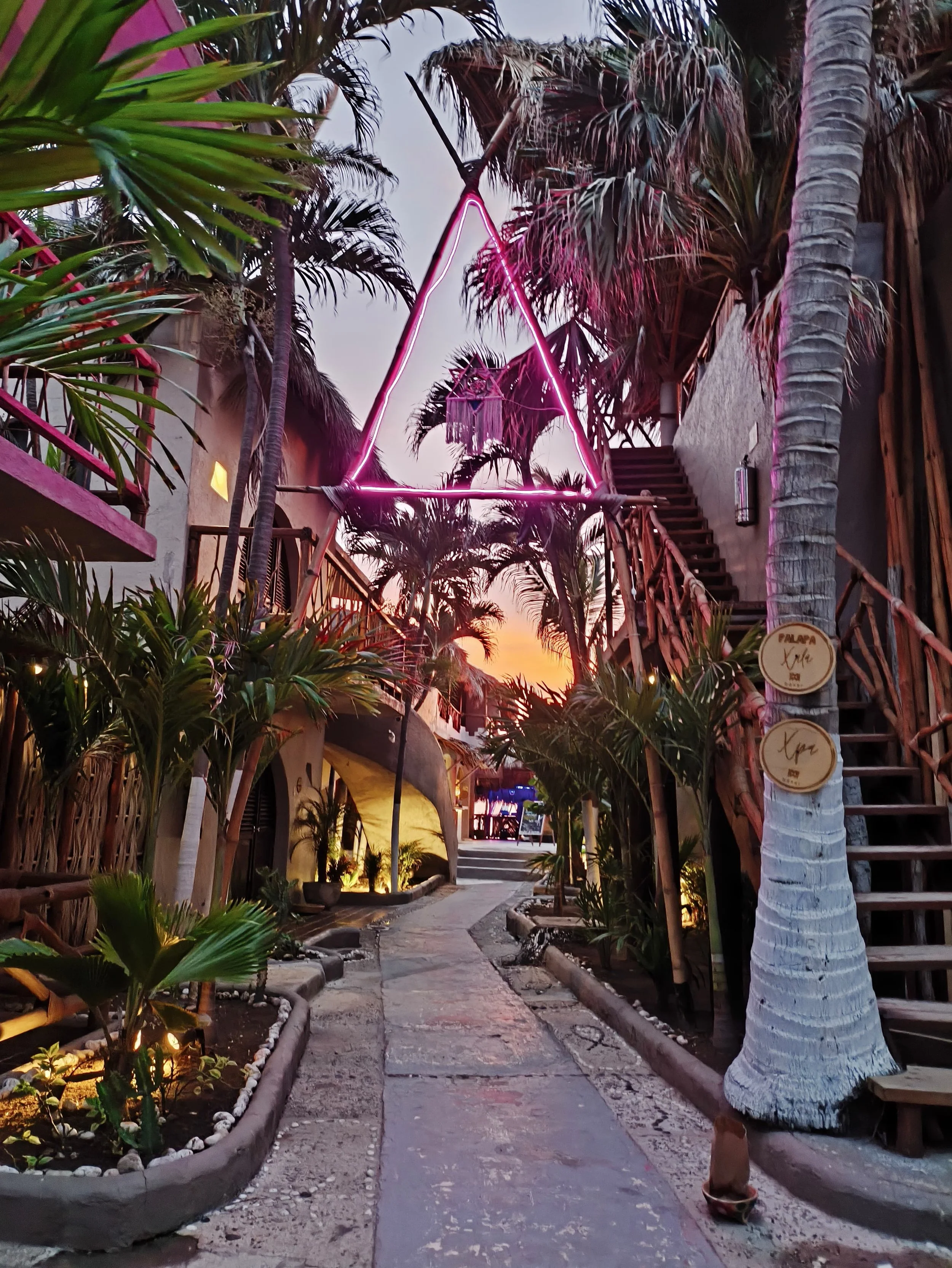 Pasillo de un espacio exterior decorado con plantas tropicales, iluminación ambiental y una estructura con neón en forma de triángulo suspendida en el aire, al atardecer. Hotel baXar pie de la cuesta puesta de sol playa pie de la cuesta