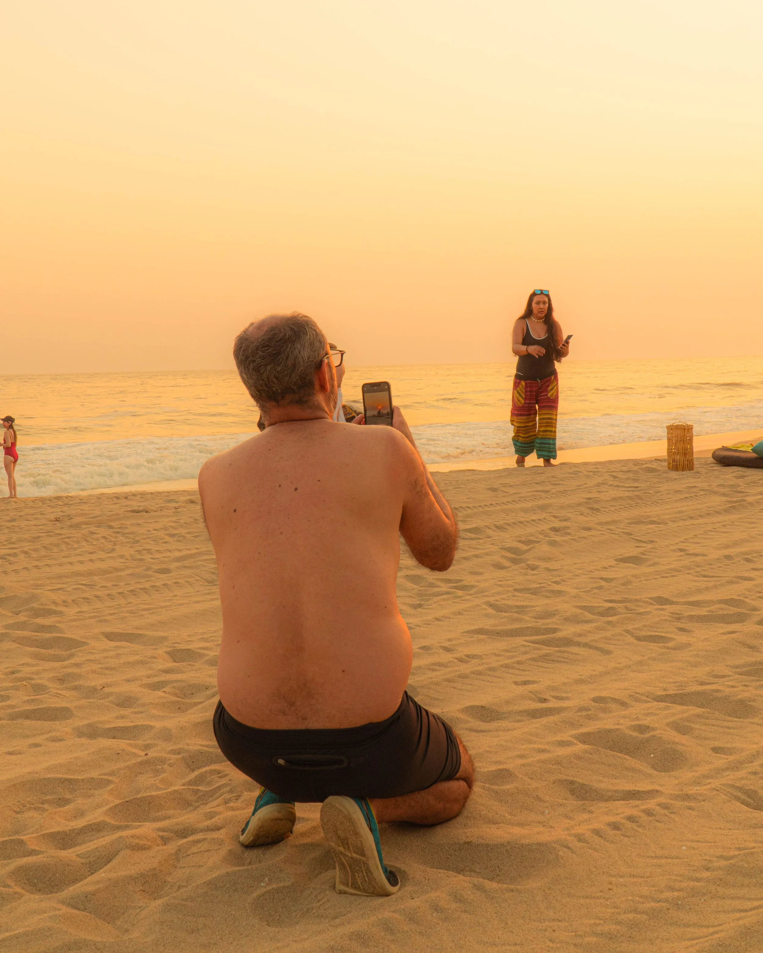 Hombre en posición de oración en la playa, con mujer de pie sosteniendo un teléfono y un paisaje de atardecer con mar en el fondo. Hotel baXar pie de la cuesta puesta de sol playa pie de la cuesta