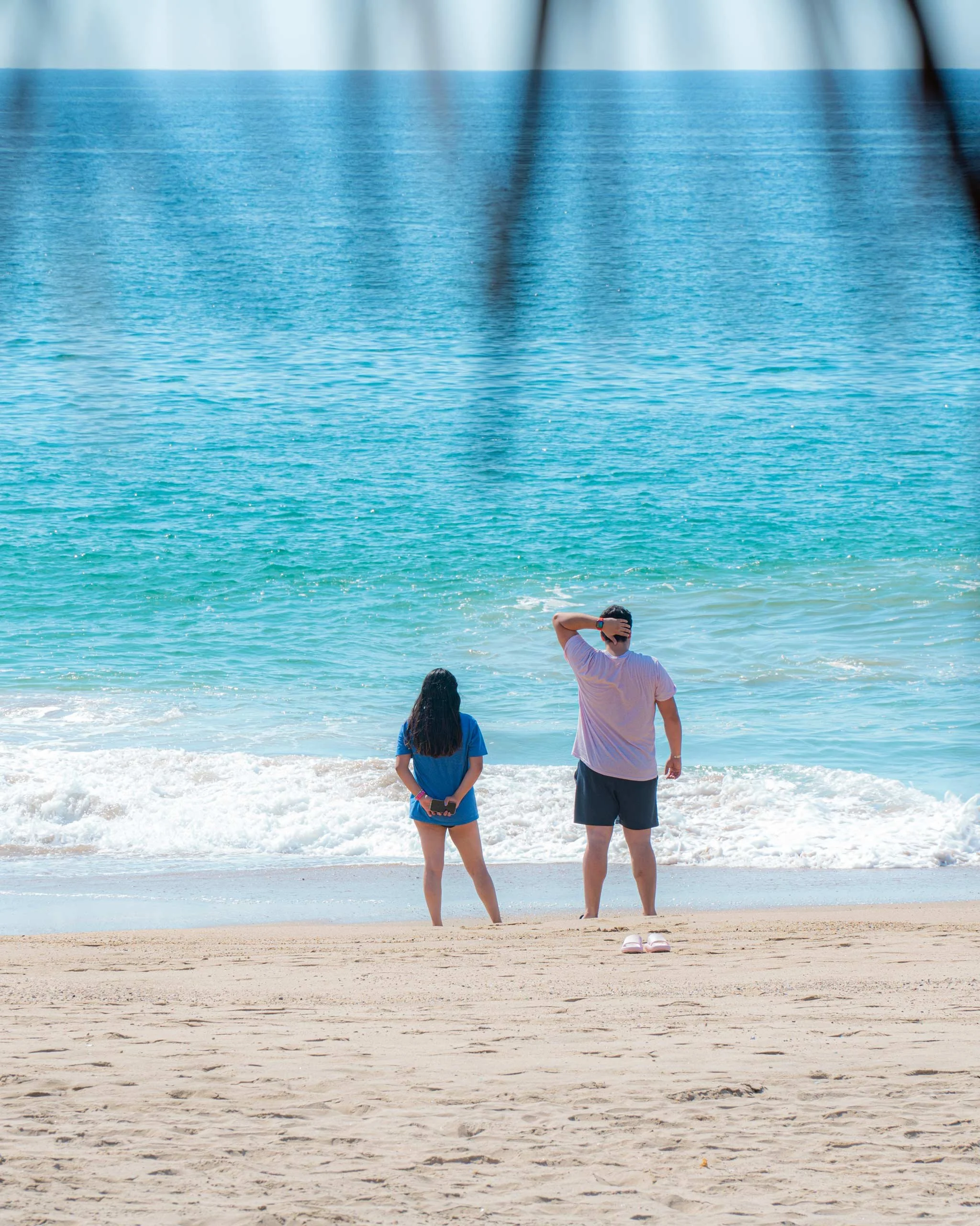 Dos personas, un niño y un adulto, observan el mar desde la orilla de la playa, con el océano extendido en el horizonte y el cielo despejado. Hotel boutique playa pie de la cuesta, baXar Acapulco