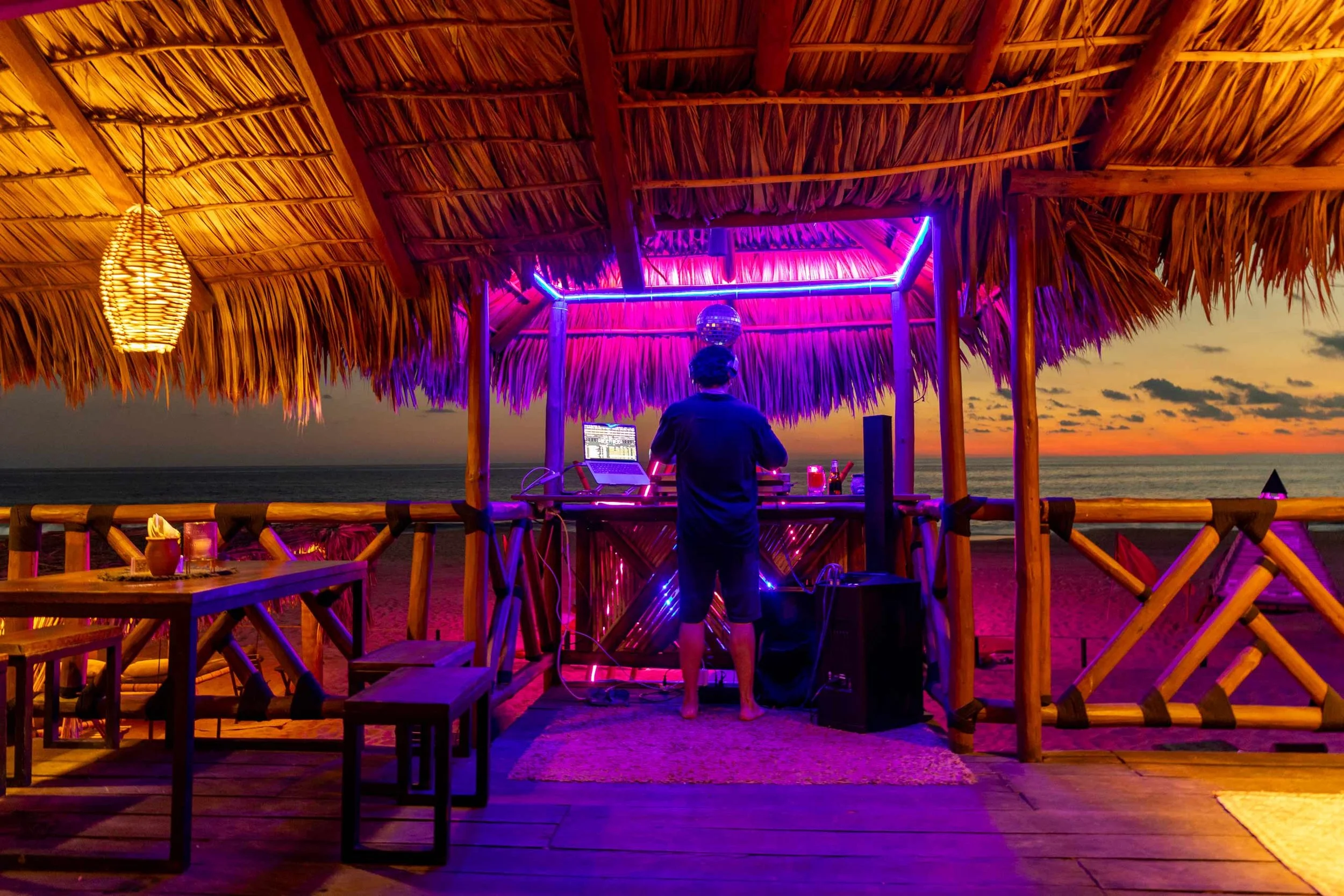 baXar Club de playa Pie de la Cuesta con techo de palma, luces de neón y un DJ tocando música en la playa al atardecer.