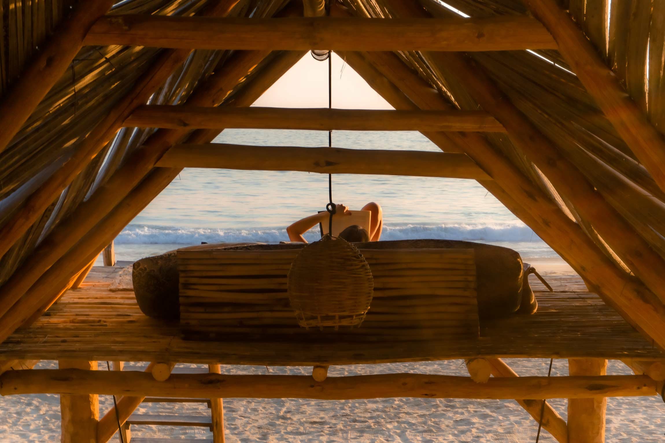Mujer acostada en una hamaca bajo una estructura de madera, viendo el mar al atardecer desde la playa en el hotel baXar pie de la cuesta