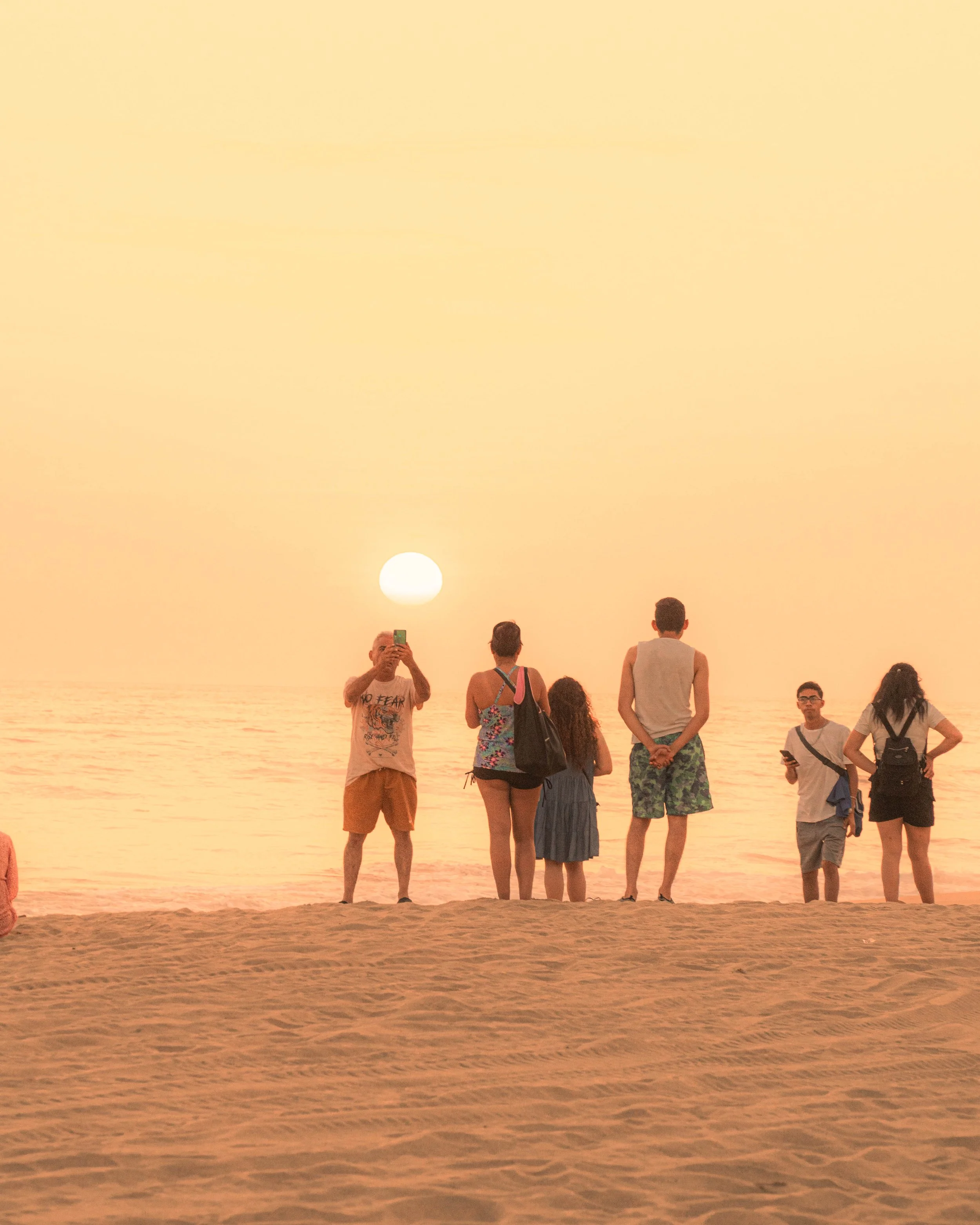 Grupo de personas en la playa al atardecer, con el sol visible en el horizonte, algunos tomando fotos y otros observando el paisaje. Hotel baXar pie de la cuesta