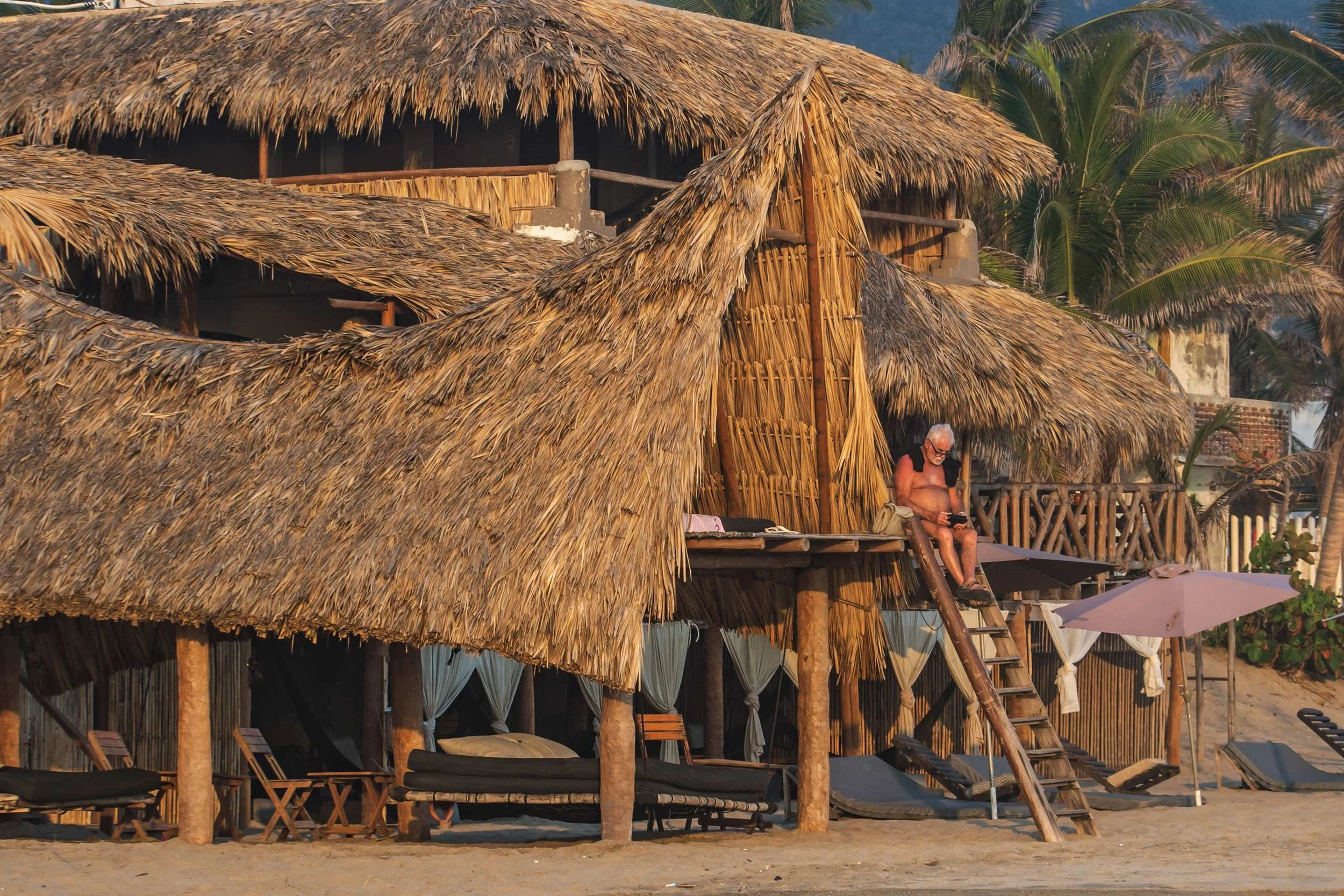 Una estructura de palapa de madera y techos de palma en una playa, con personas descansando y palmeras de fondo. day pass beach club de playa pie de la cuesta, hotel boutique baXar acapulco