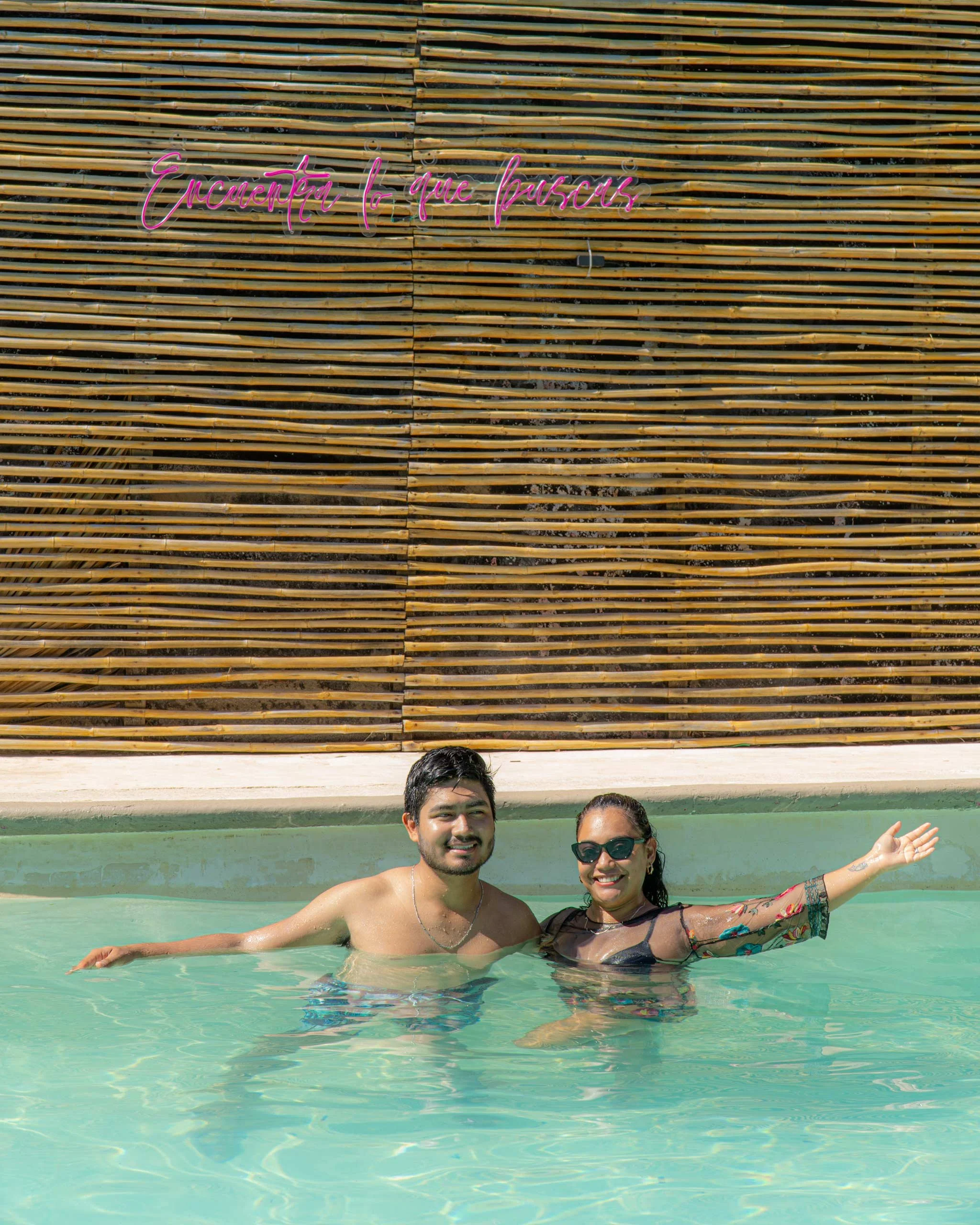 Dos personas sonriendo y usando trajes de baño, nadando en una piscina de agua clara, con una pared de madera en el fondo y un cartel con escritura en neón que dice 'Encuentra lo que buscas'.  club de playa pie de la cuesta, hotel boutique baXar