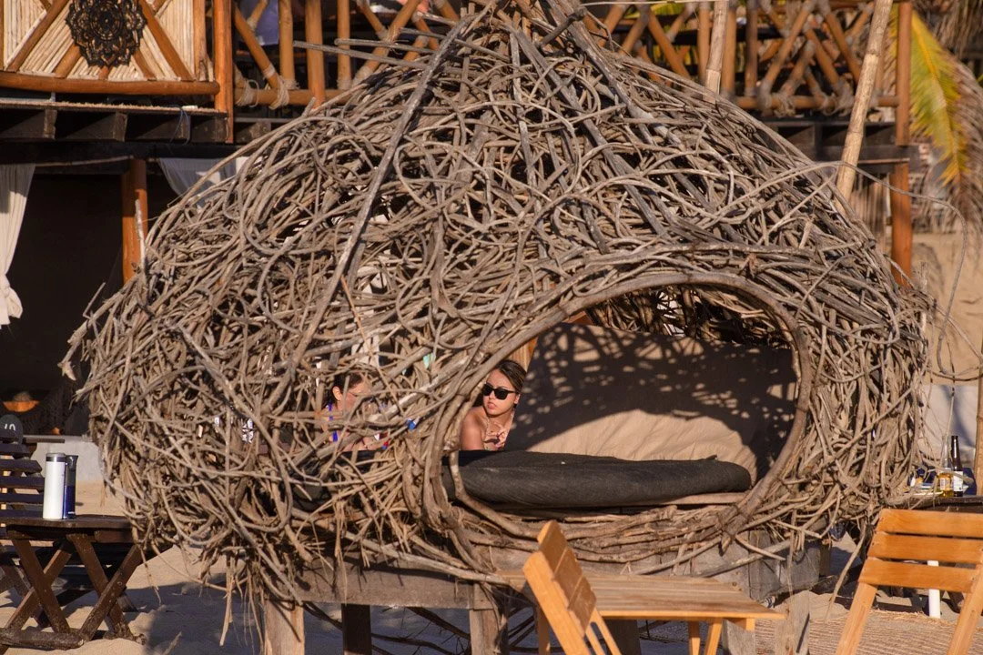 Estructura de mimbre en forma de cabaña en una playa con personas sentadas debajo, con sombra y muebles de madera. club de playa pie de la cuesta, hotel boutique baXar acapulco,