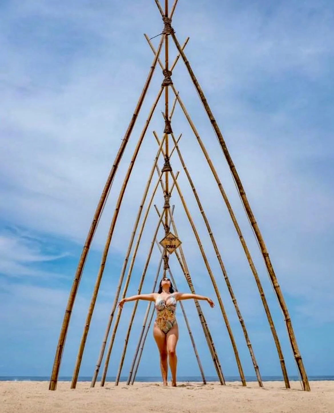 Mujer con traje de baño en la playa, posando debajo de una estructura de varas de madera. La estructura forma un arco grande con un aviso de advertencia en el centro. El cielo está despejado y húmedo. Hotel baXar pie de la cuesta puesta de sol playa 