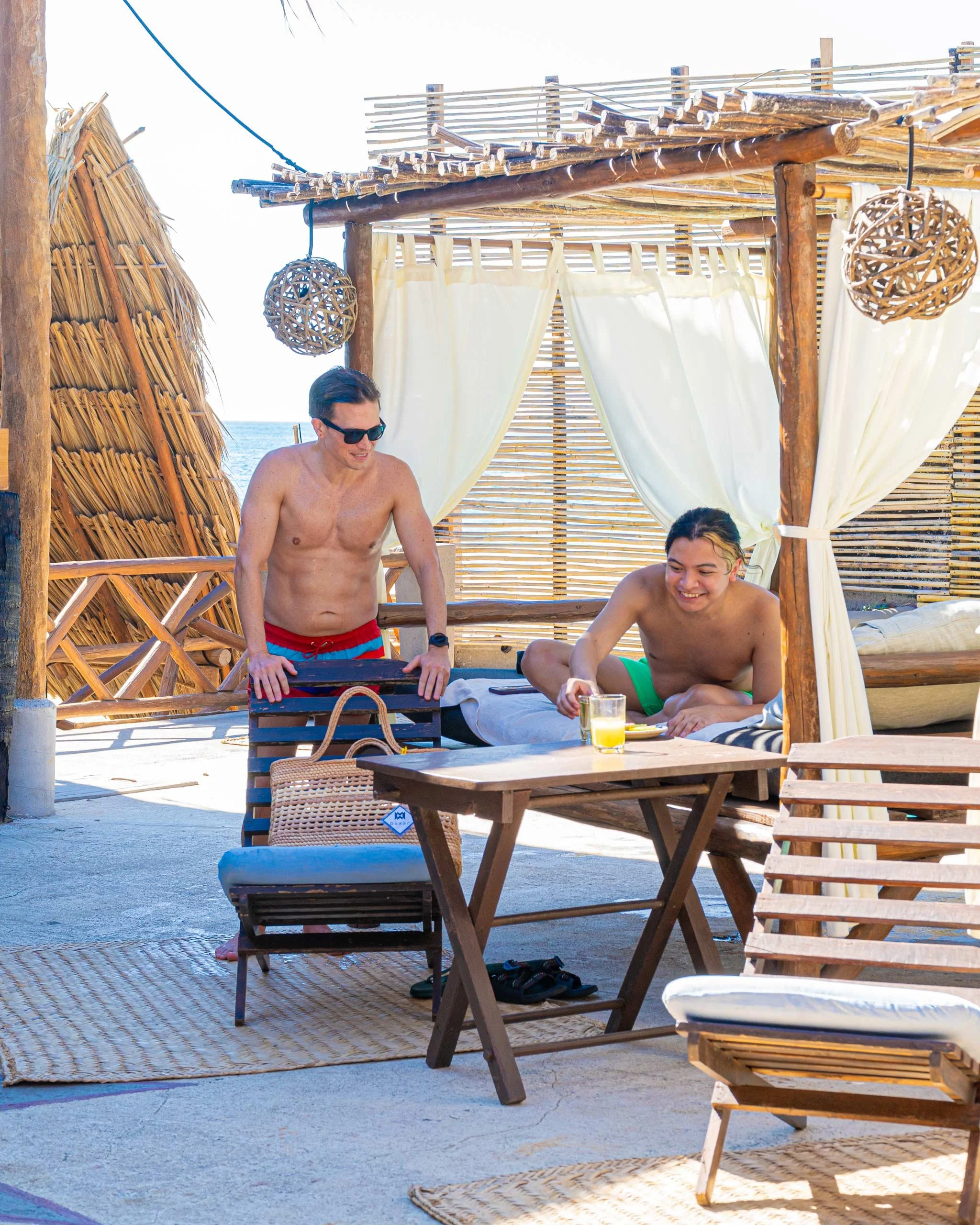 Dos hombres en trajes de baño en una estructura de playa con cortinas blancas, lámparas de mimbre y vista al mar en el fondo. Hotel boutique baXar playa Pie de la cuesta Acapulco