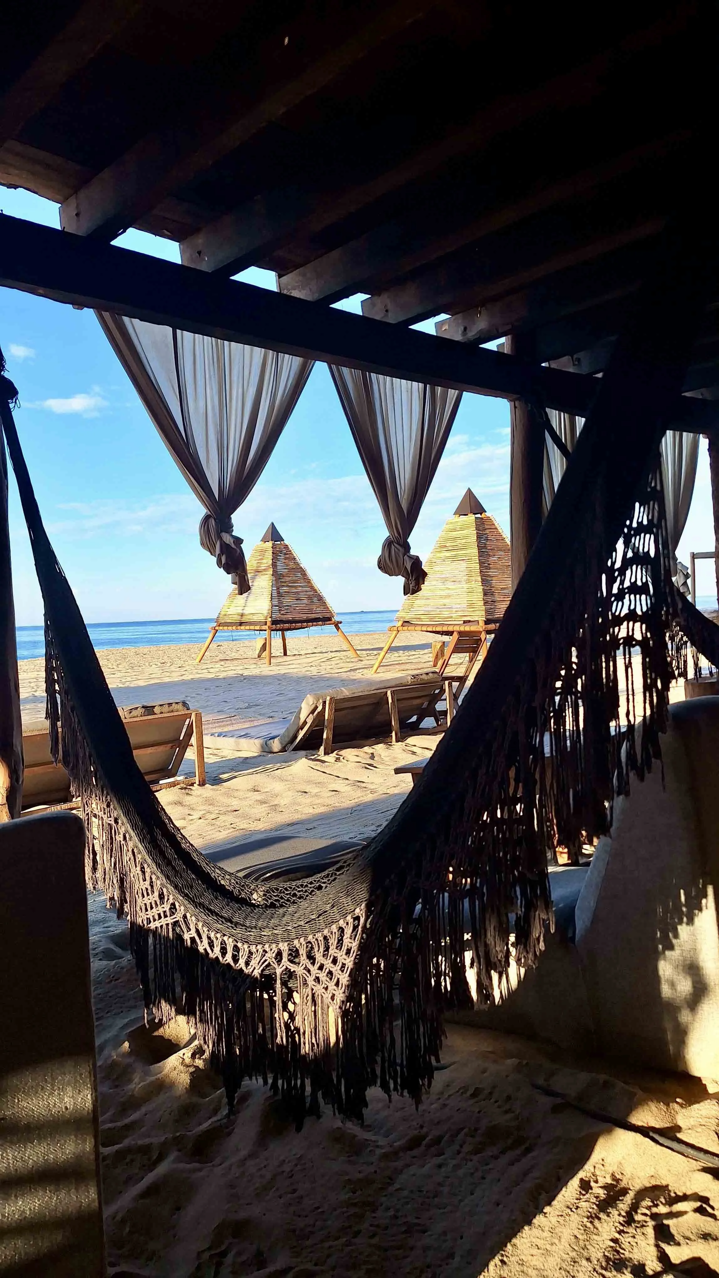 Hamaca oscura en un espacio de descanso en la playa con vistas al mar y cabañas de techo de paja al fondo bajo un cielo despejado.