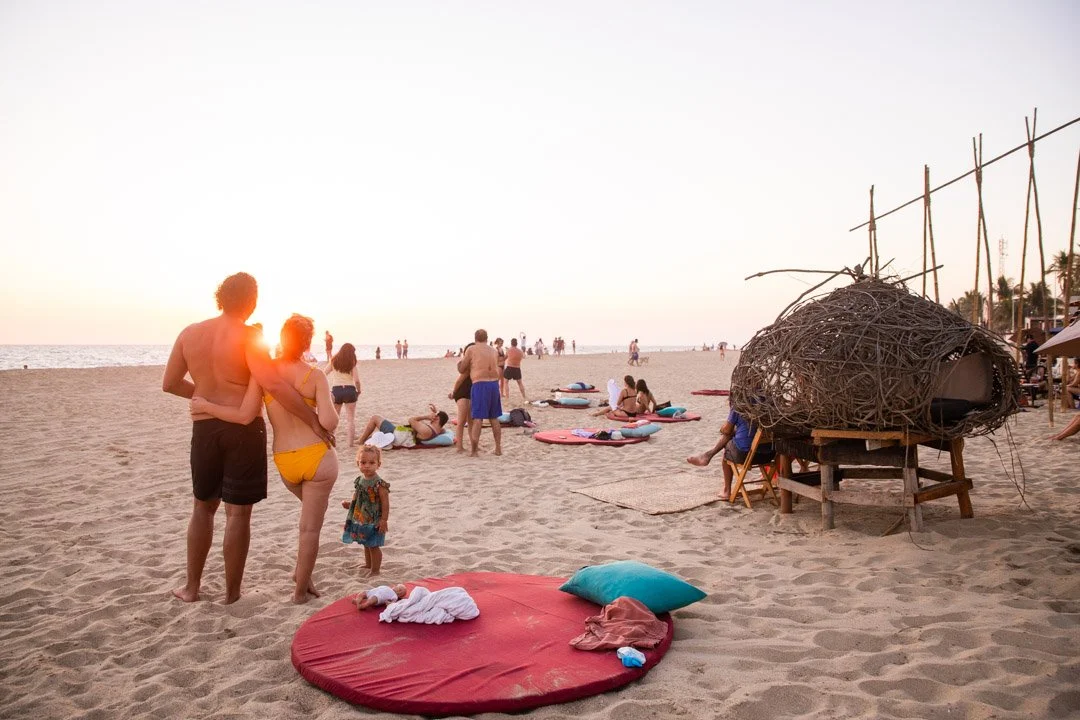 Familia en la playa al atardecer, con personas descansando y jugando sobre la arena, y una estructura de madera con enredaderas a la derecha. beach club day pass club de playa pie de la cuesta, hotel boutique baXar acapulco