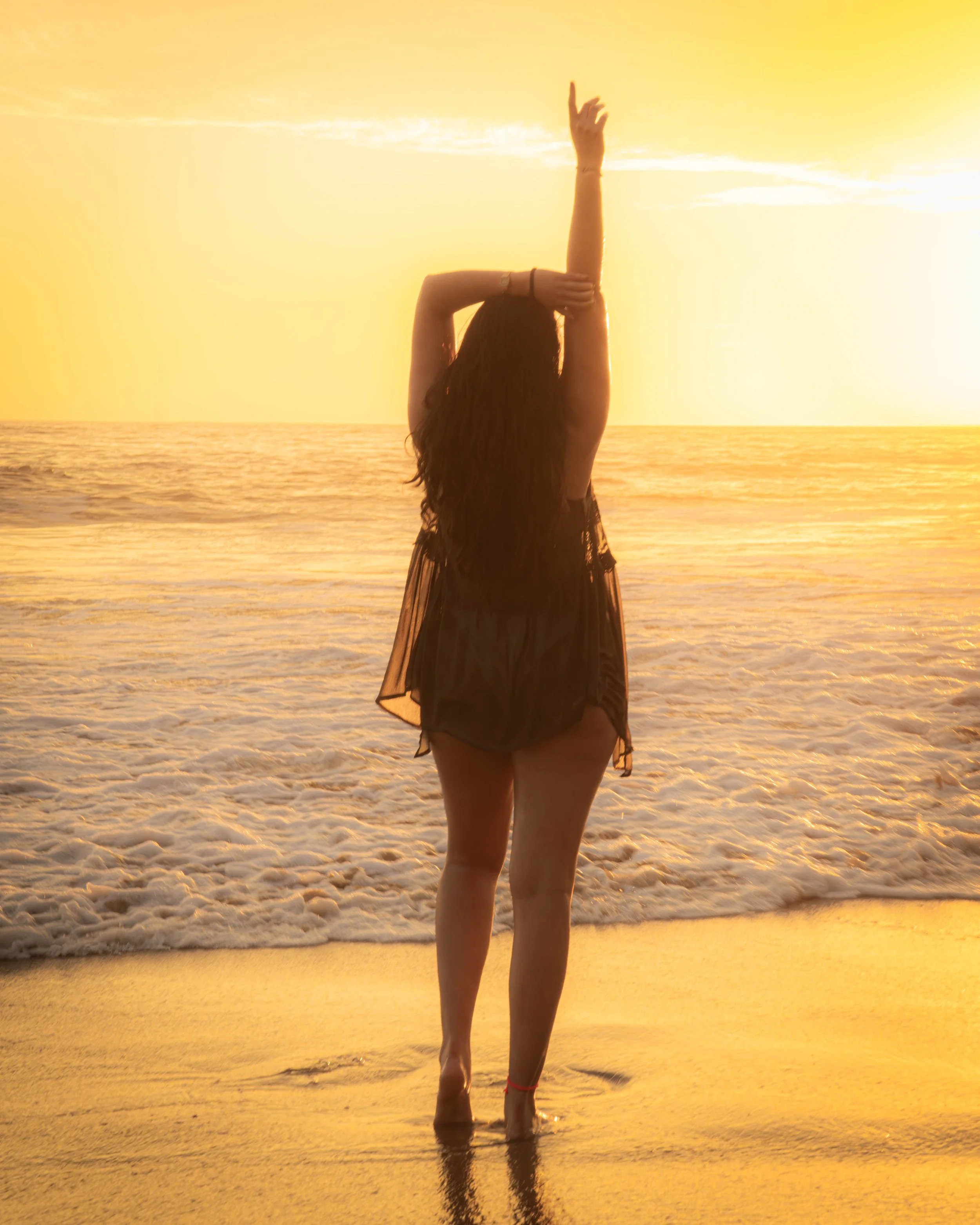 Mujer de espaldas en la orilla del mar durante una puesta de sol, con vestido negro y brazos en posición de stretching.  playa Pie de la cuesta, Acapulco, Hotel boutique baXar 