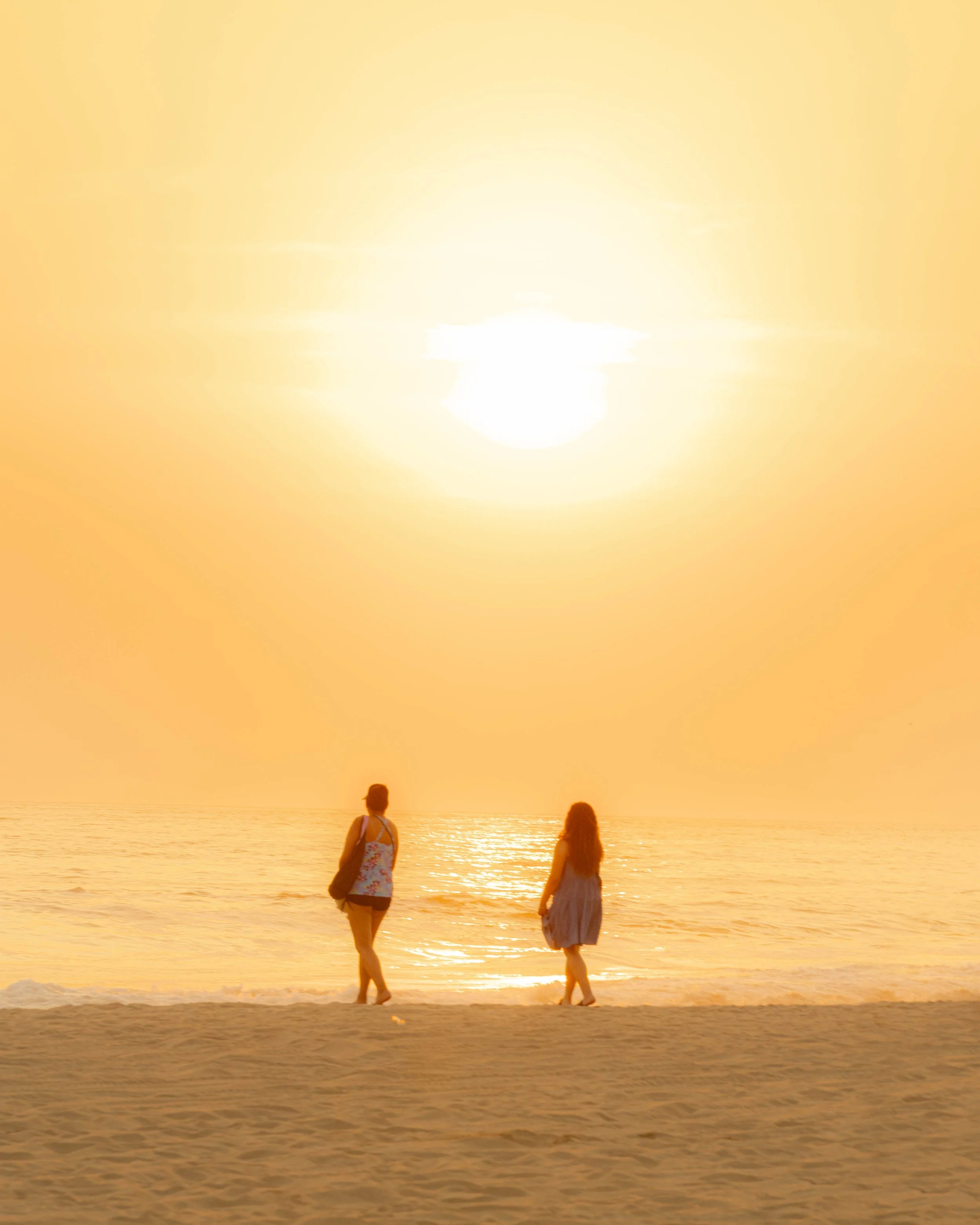 Dos mujeres caminando por la playa pie de la cuesta durante un atardecer, con el sol en el horizonte y el mar a sus lados. baxar hotel y club de playa acapulco