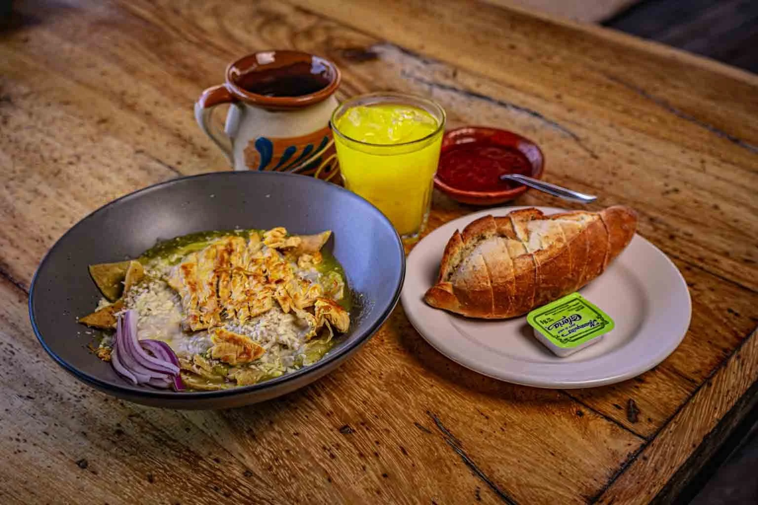 Plato de huevos con tortilla, plátano, y tomate, pan, y bebidas con café y jugo en una mesa de madera.
