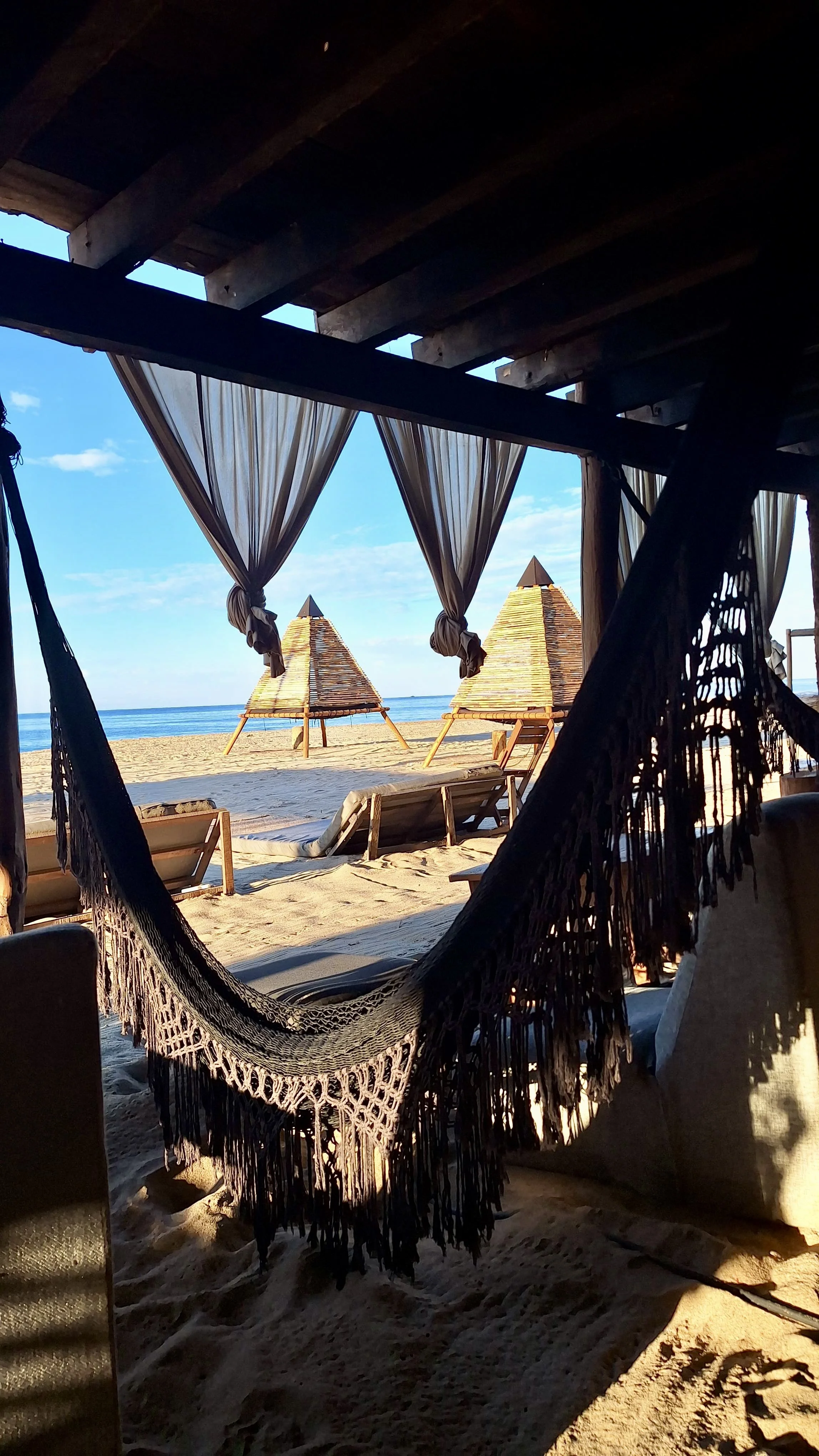 Hamaca colgando en una cabaña con vista a la playa, con sombrillas de madera en la arena, al fondo se ve el mar y el cielo despejado.  beach club day pass club de playa pie de la cuesta, hotel boutique baXar acapulco