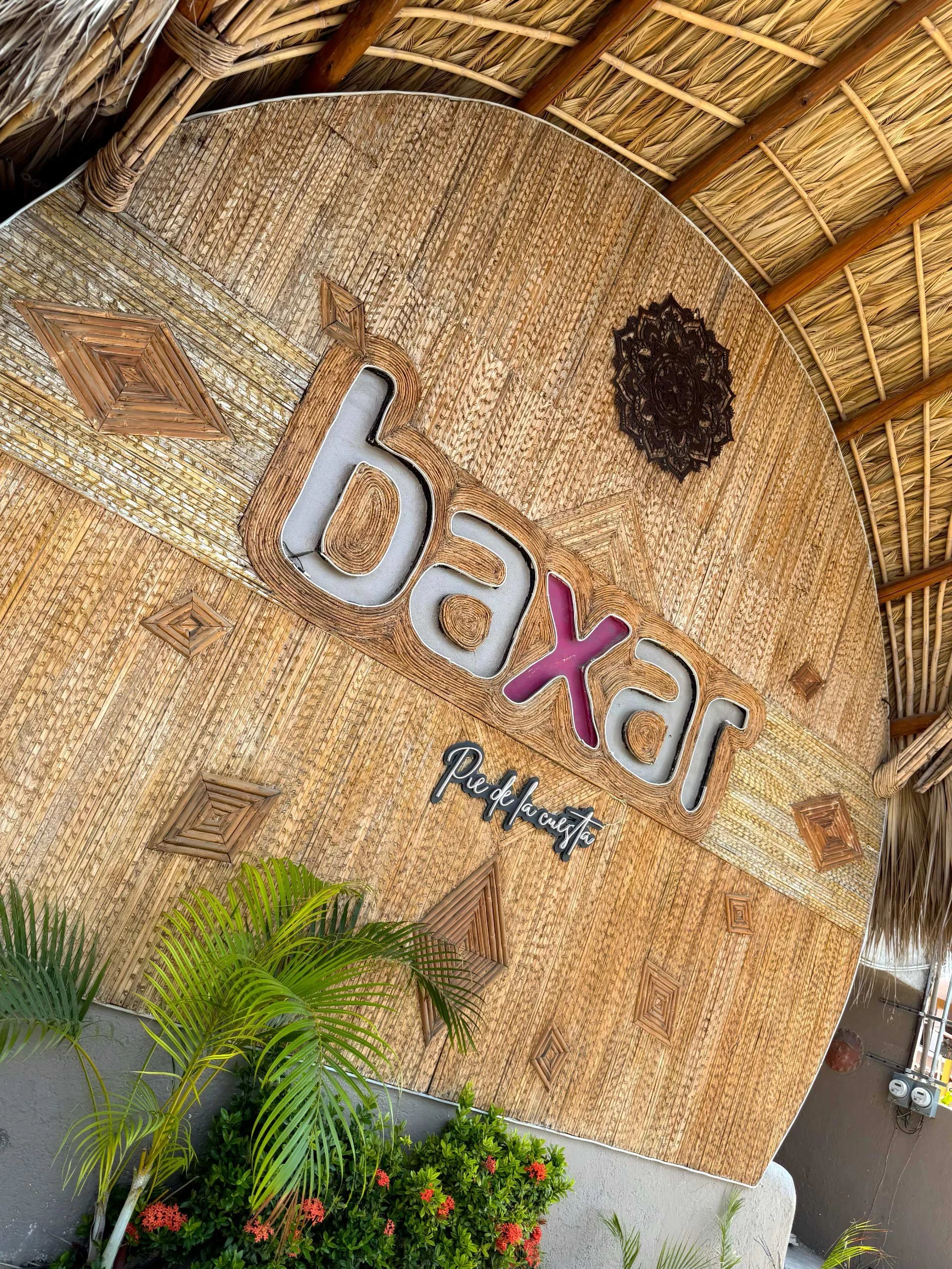 Se observa un cartel con letras grandes que dice 'BOX', en un entorno con decoración de estilo rústico, con paredes de madera y techo de paja. Hay plantas verdes y flores rojas en la parte inferior de la imagen. day pass beach club