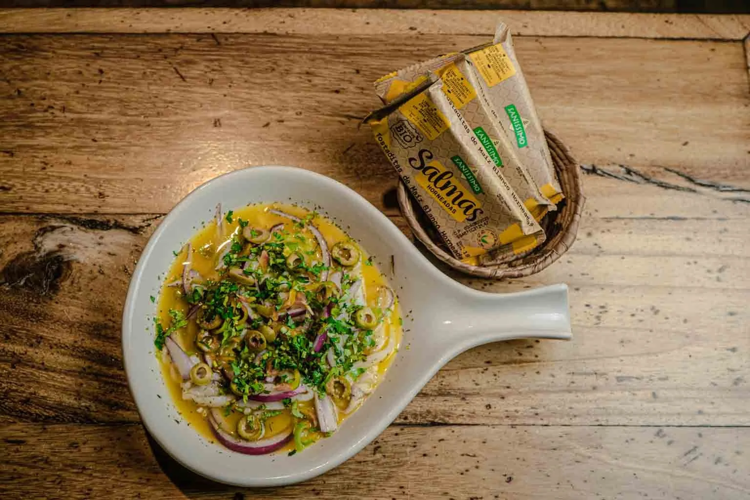 En un plato de cerámica en forma de pala hay ensalada de cebolla, cilantro y chiles en vinagre y guacamole. Al lado, en un canasto de mimbre, hay un paquete de salmorejo salmón.