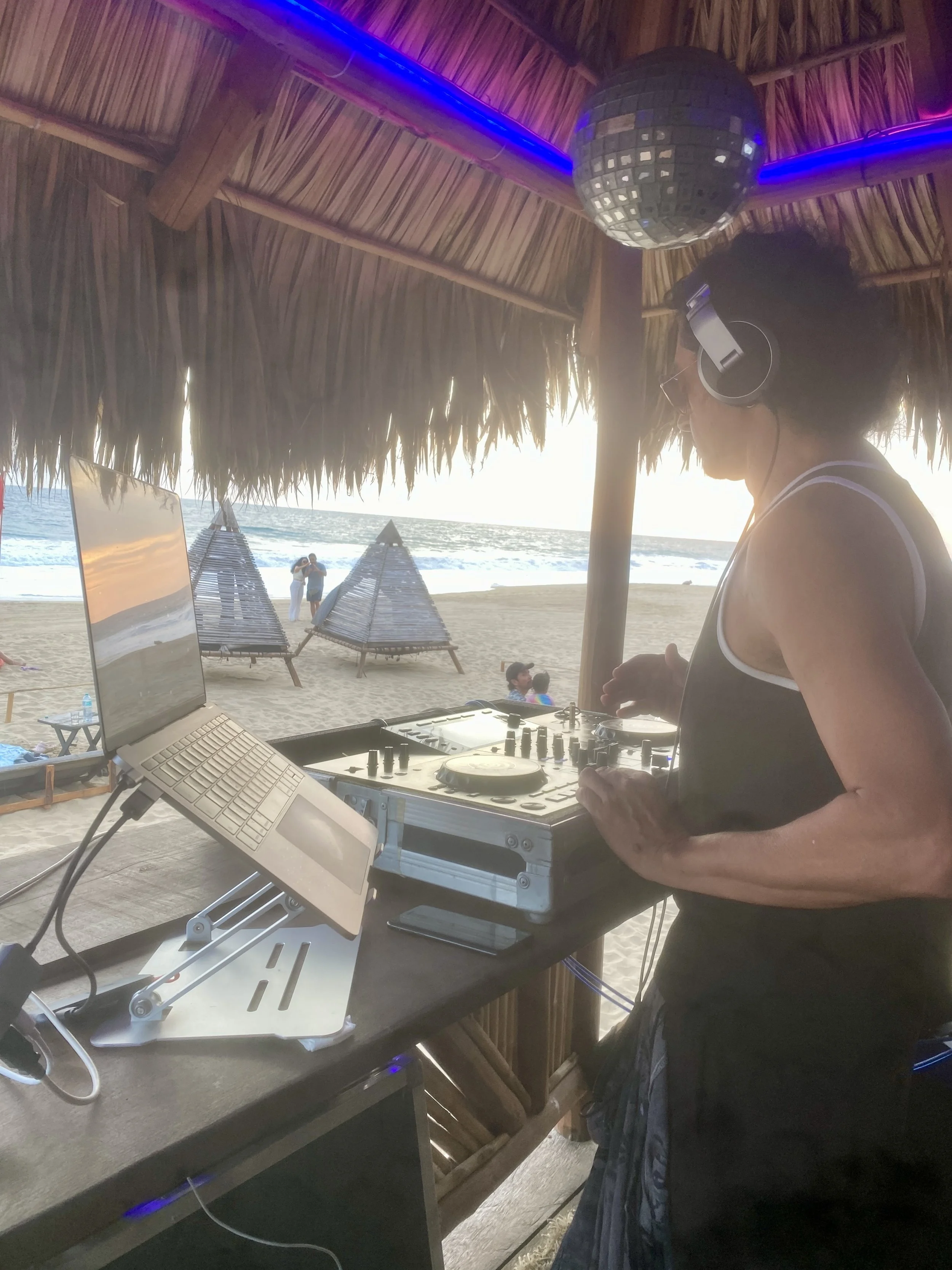 DJ JABA mezclando música en un equipo de DJ en baXar Playa Pie de la Cuesta, con palapas y sillas de playa, y un espejo que refleja la escena y la playa al atardecer.