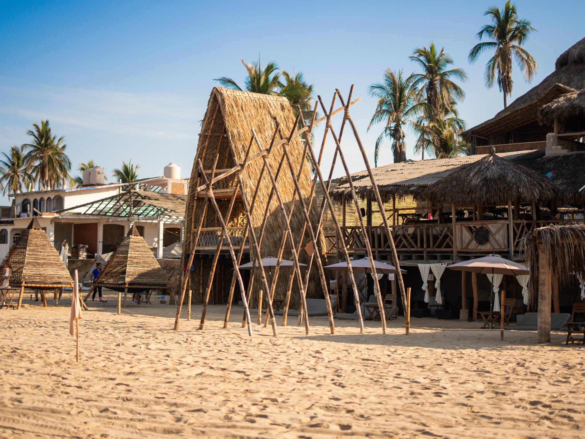 Estructuras de madera y techos de paja en una playa, con palmeras y cielo azul al fondo. day pass beach club de playa pie de la cuesta, hotel boutique baXar acapulco