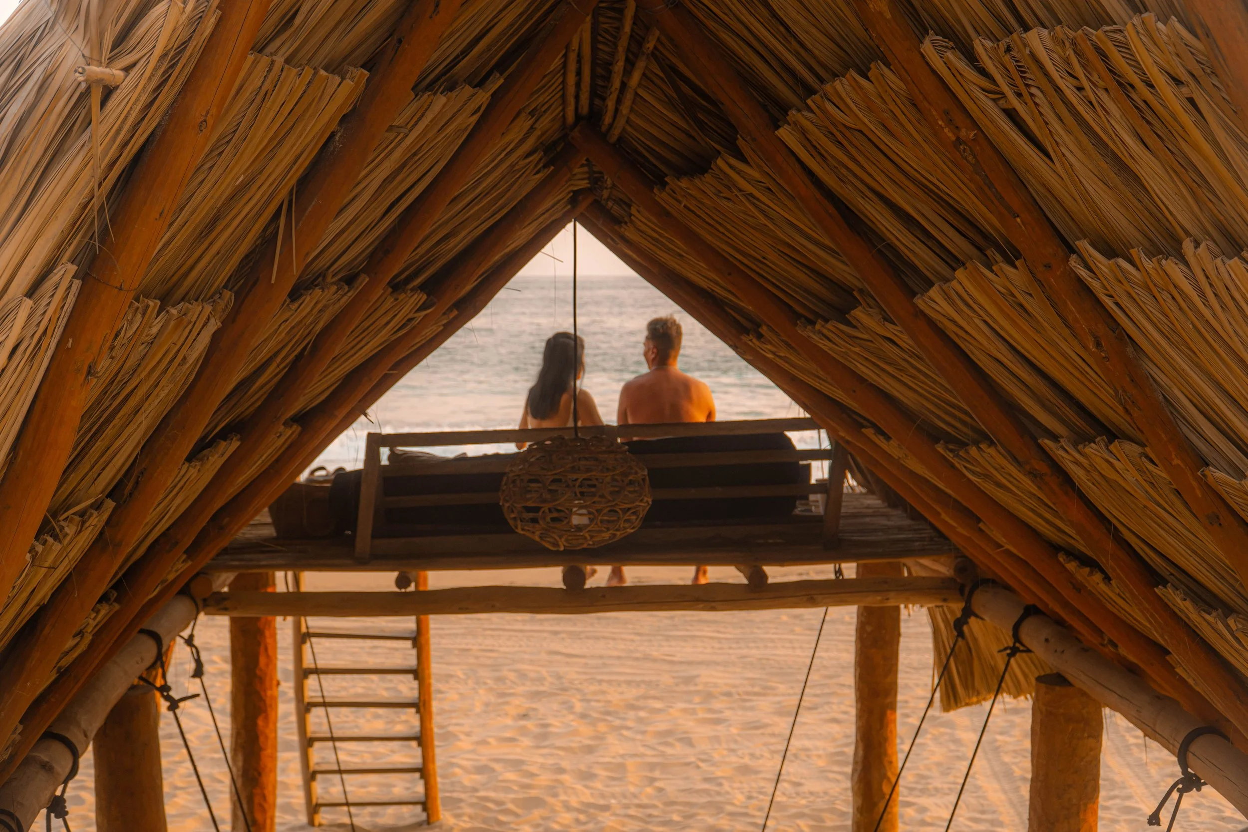 Vista desde dentro de una choza de palma en la playa, con dos personas sentadas en una banca viendo el mar durante el atardecer. day pass beach club de playa pie de la cuesta, hotel boutique baXar acapulco