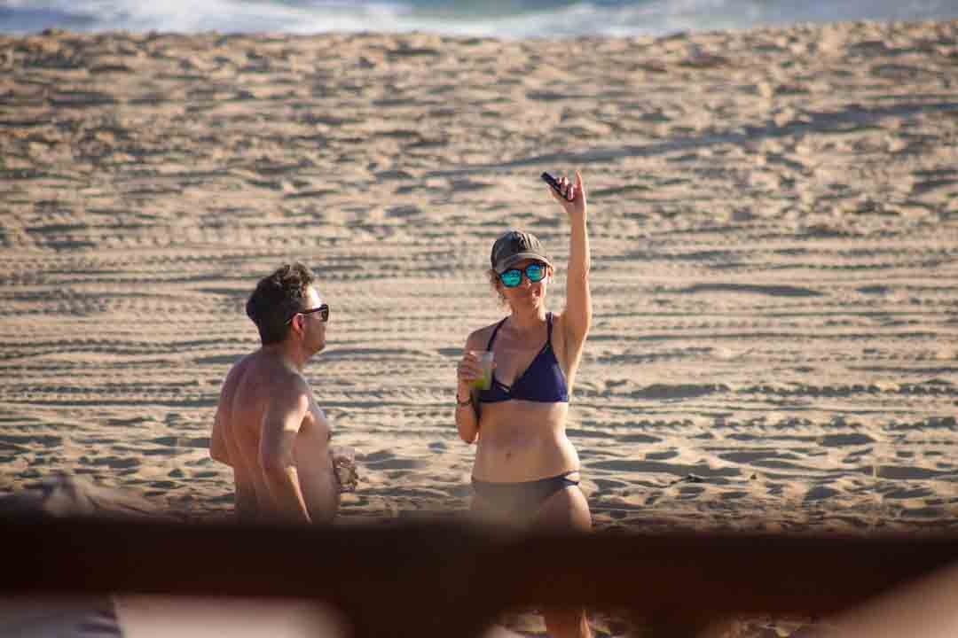 Dos personas en la playa, una mujer con traje de baño y gafas de sol levantando la mano, un hombre sin camisa con gafas de sol, ambos en un ambiente soleado de arena. hotel baxar pie de la cuesta