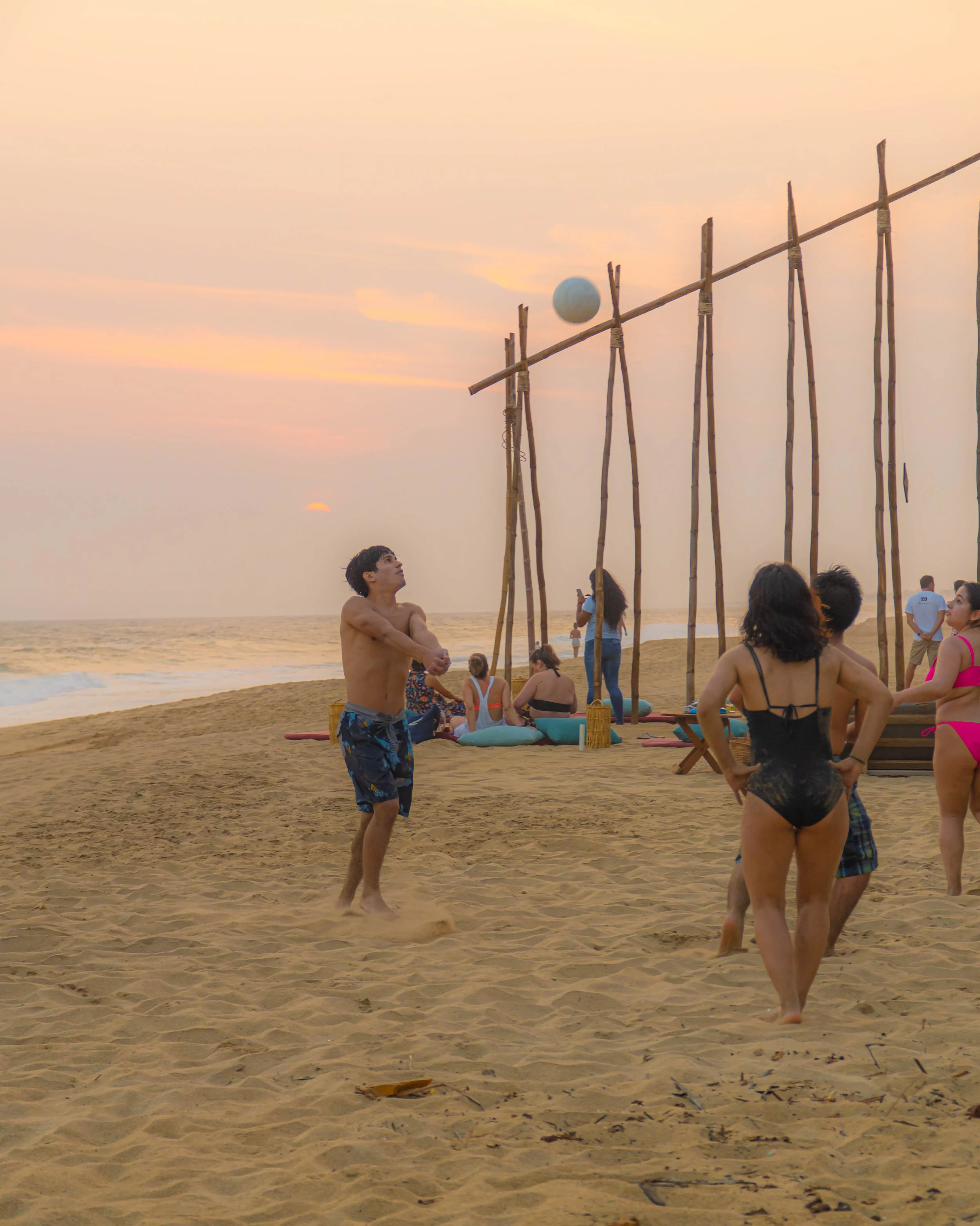 Personas en la playa participando en una actividad de voley playa con un fuerte y una estructura de madera, durante el atardecer. club de playa pie de la cuesta, beach club day pass hotel baxar