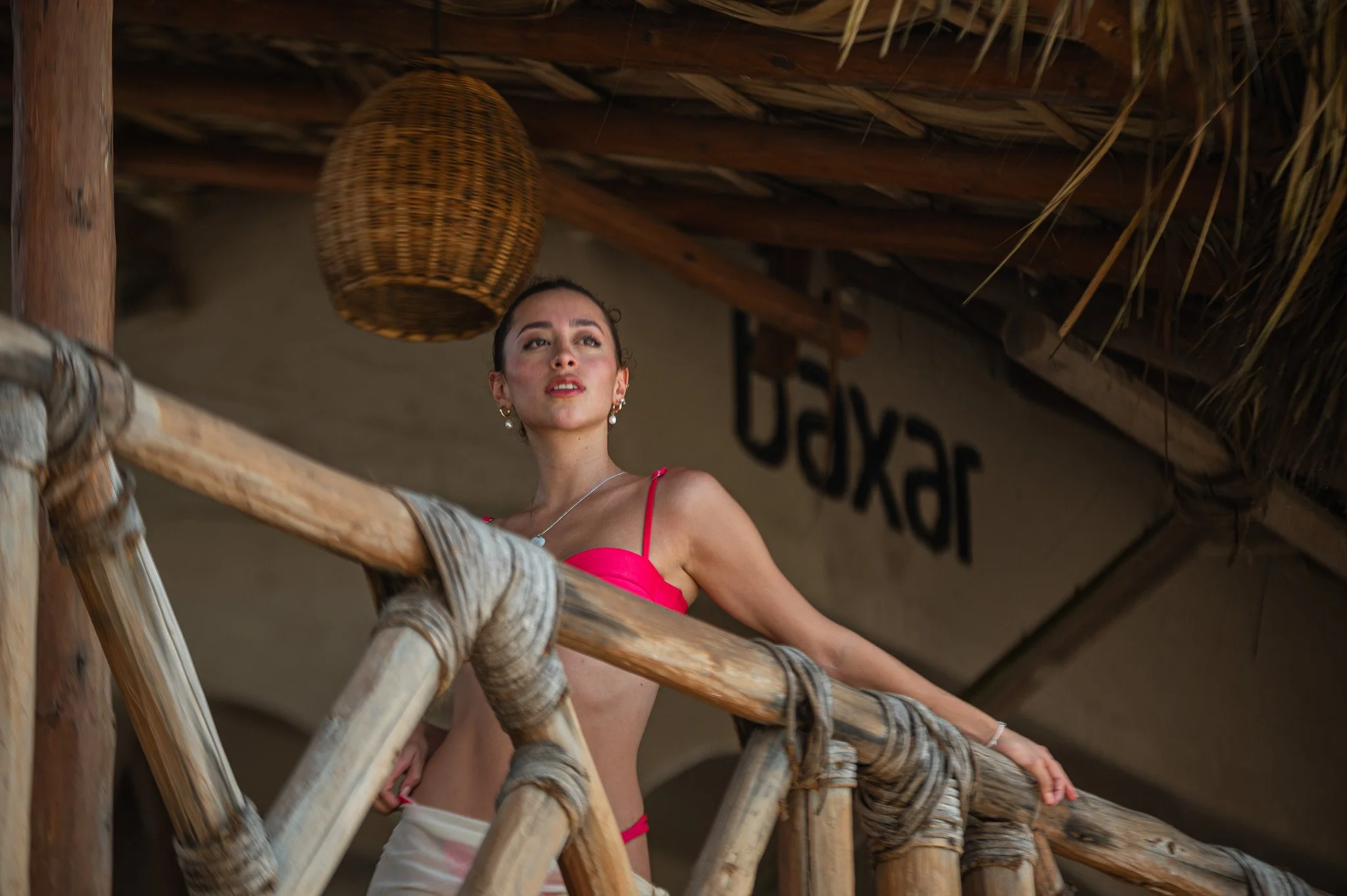 Mujer joven en trajes de baño rosa, en una estructura de madera en un entorno rústico con techo de paja.  Hotel baXar Playa de la cuesta