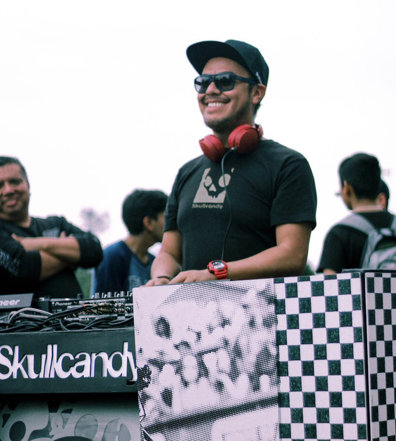 Dj Shine sonriente usando gafas de sol, gorra y audífonos rojos, mezclando música en un evento al aire libre con varias personas en el fondo.