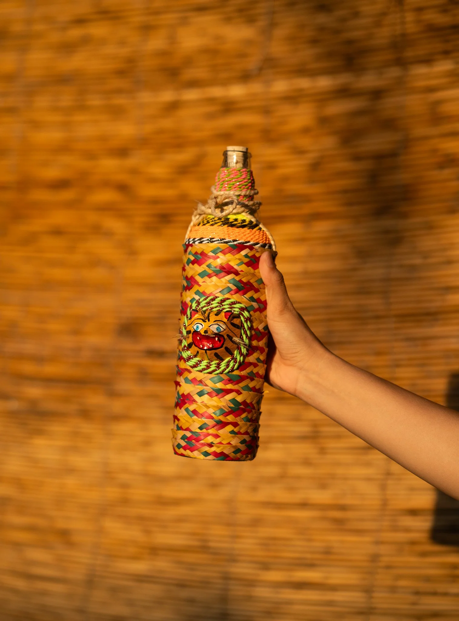 Mano sosteniendo una botella decorada con cuerda de colores y una cara de payaso pintada en ella, con fondo borroso de color naranja y marrón. Hotel baXar pie de la cuesta puesta de sol playa pie de la cuesta