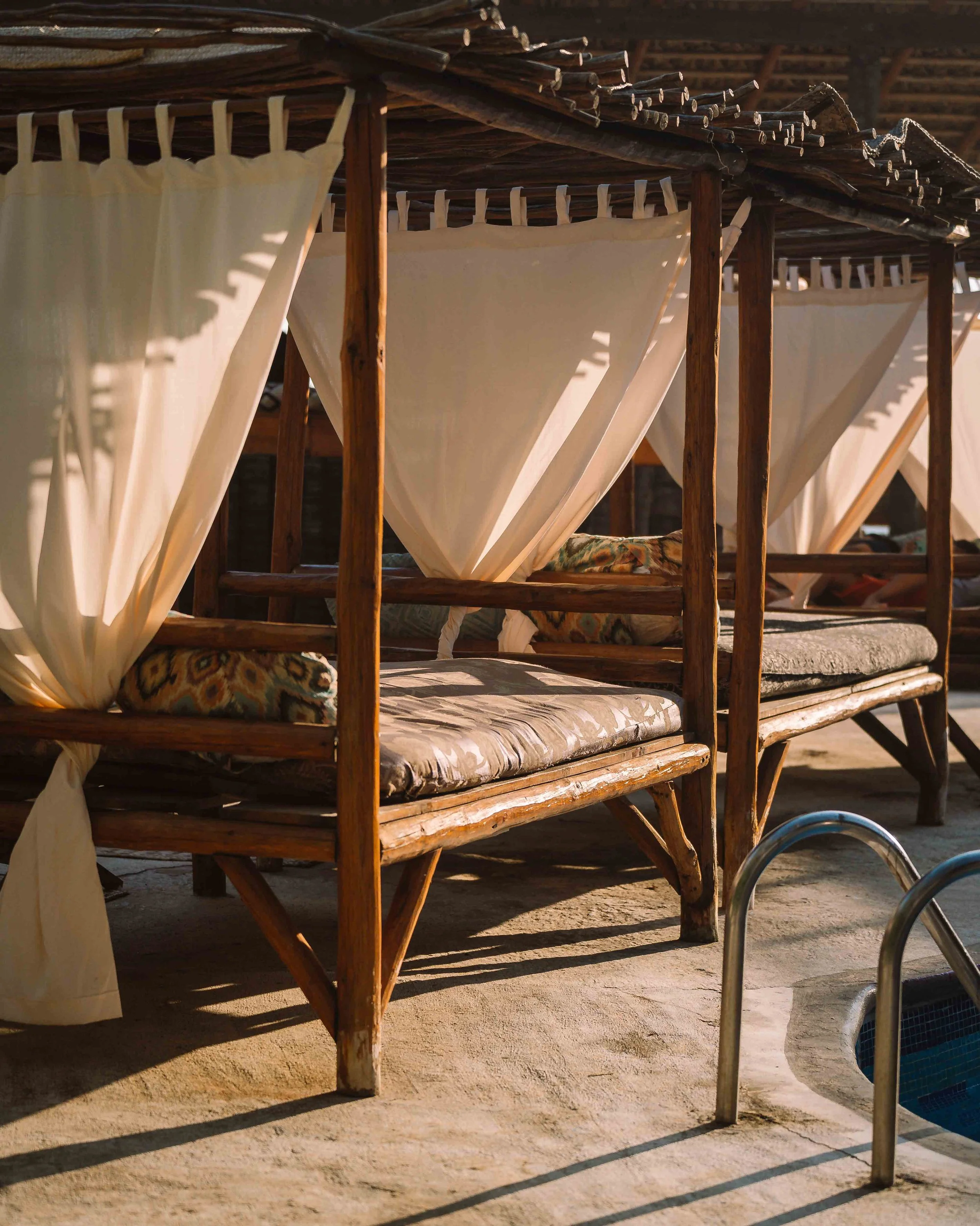 Camastros de madera con cortinas blancas cerca de la piscina, en un entorno de playa o resort. hotel baxar pie de la cuesta 