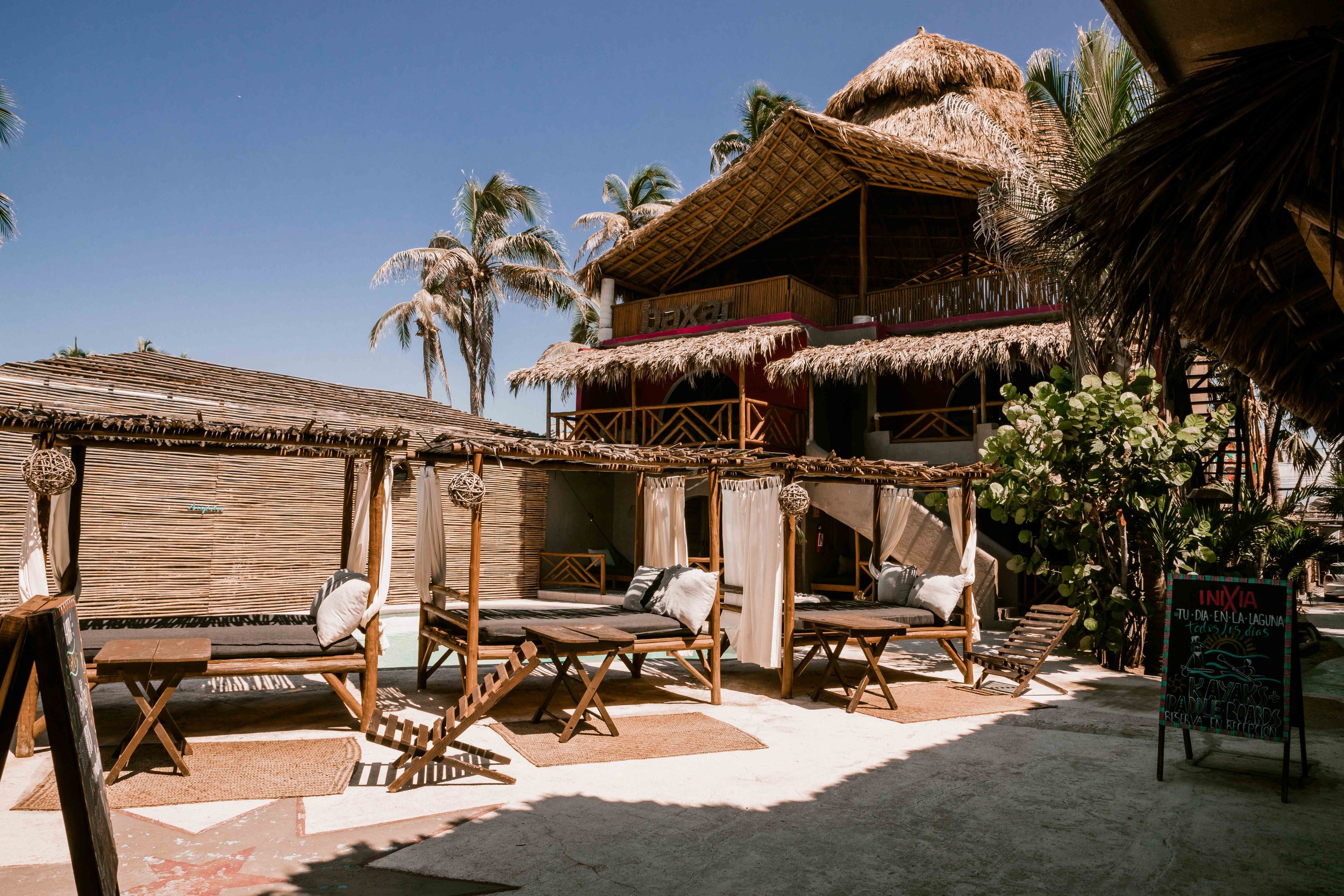 Áreas de descanso con cabañas de madera y cortinas en una playa tropical, palmeras y cielo despejado. hotel baxar pie de la cuesta 