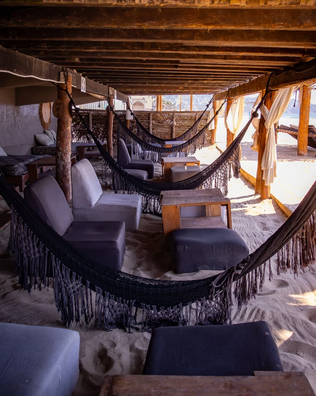 Área de descanso en una cabaña de playa, con sillones, hamacas y mesas de madera, arena en el suelo y cortinas asoleadas. day pass beach club de playa pie de la cuesta, hotel boutique baXar acapulco