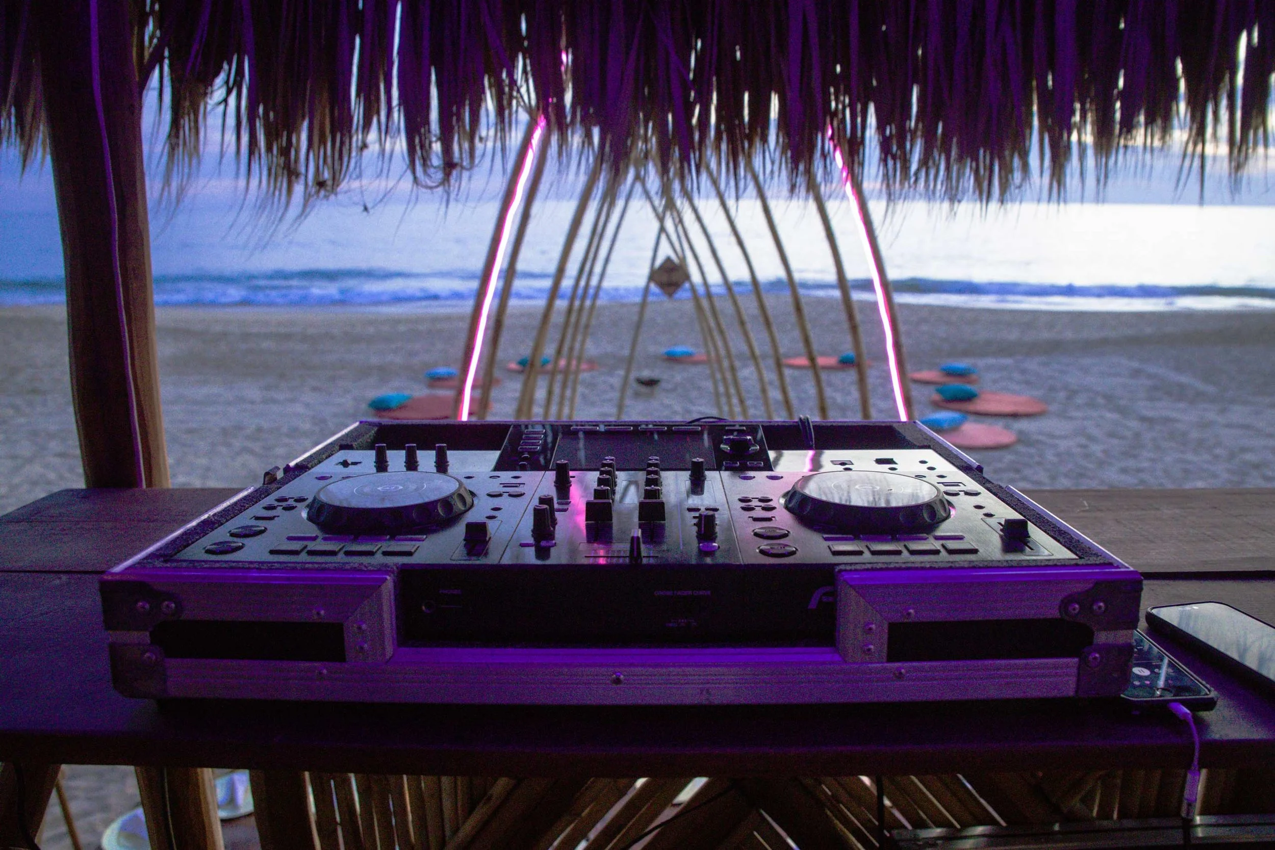 Configurador de DJ en una estructura de madera, con vista a la playa y al mar, con una palapa de paja y luces color rosa en el atardecer. baXar music - Playa pie de la cuesta, Acapulco, Mexico