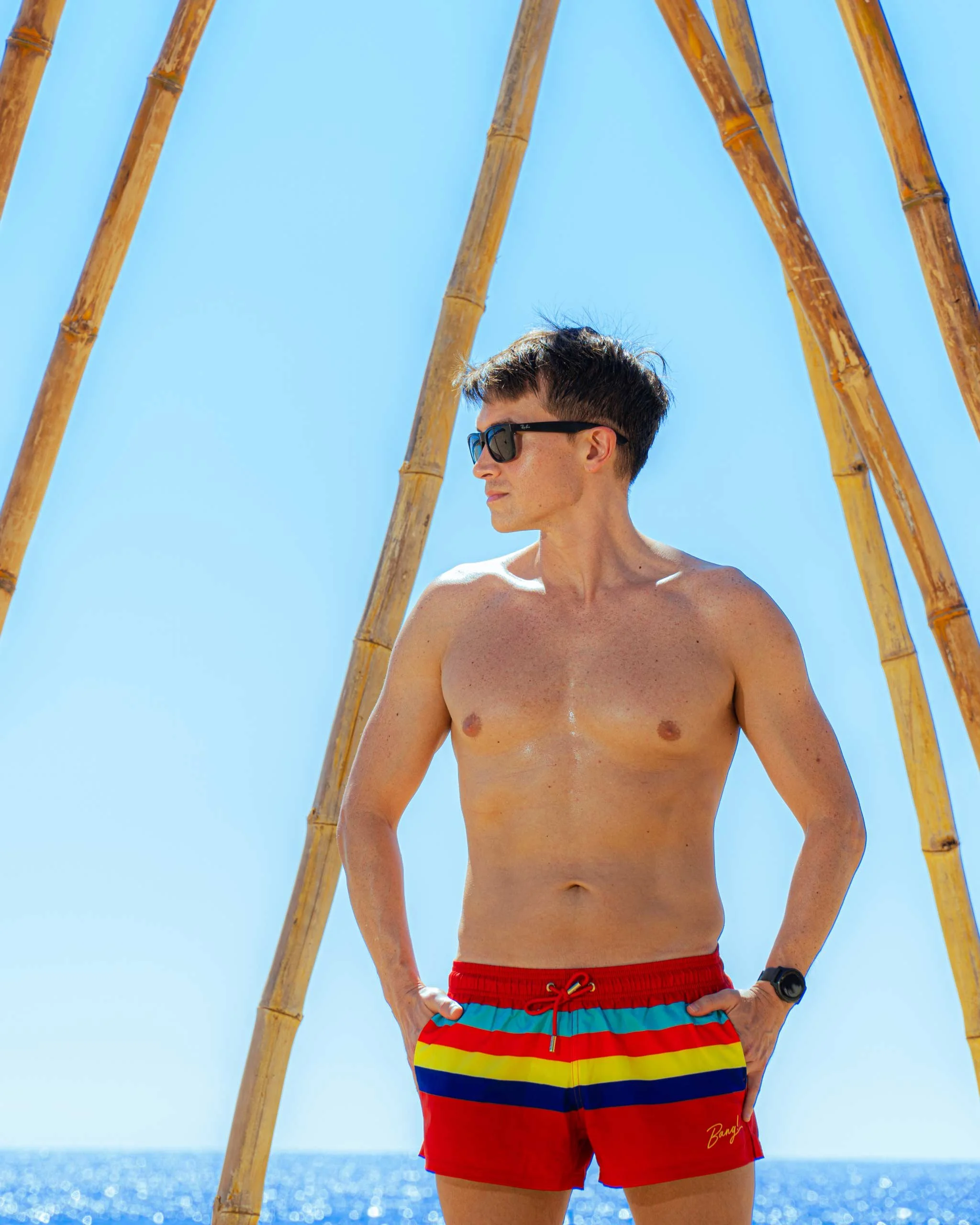 Hombre de cabello corto, usando gafas de sol, sin camiseta, con pantalones cortos a rayas de colores, de pie frente a estructuras de bambú en la playa con mar y cielo azul de fondo. Hotel boutique baXar playa Pie de la cuesta Acapulco