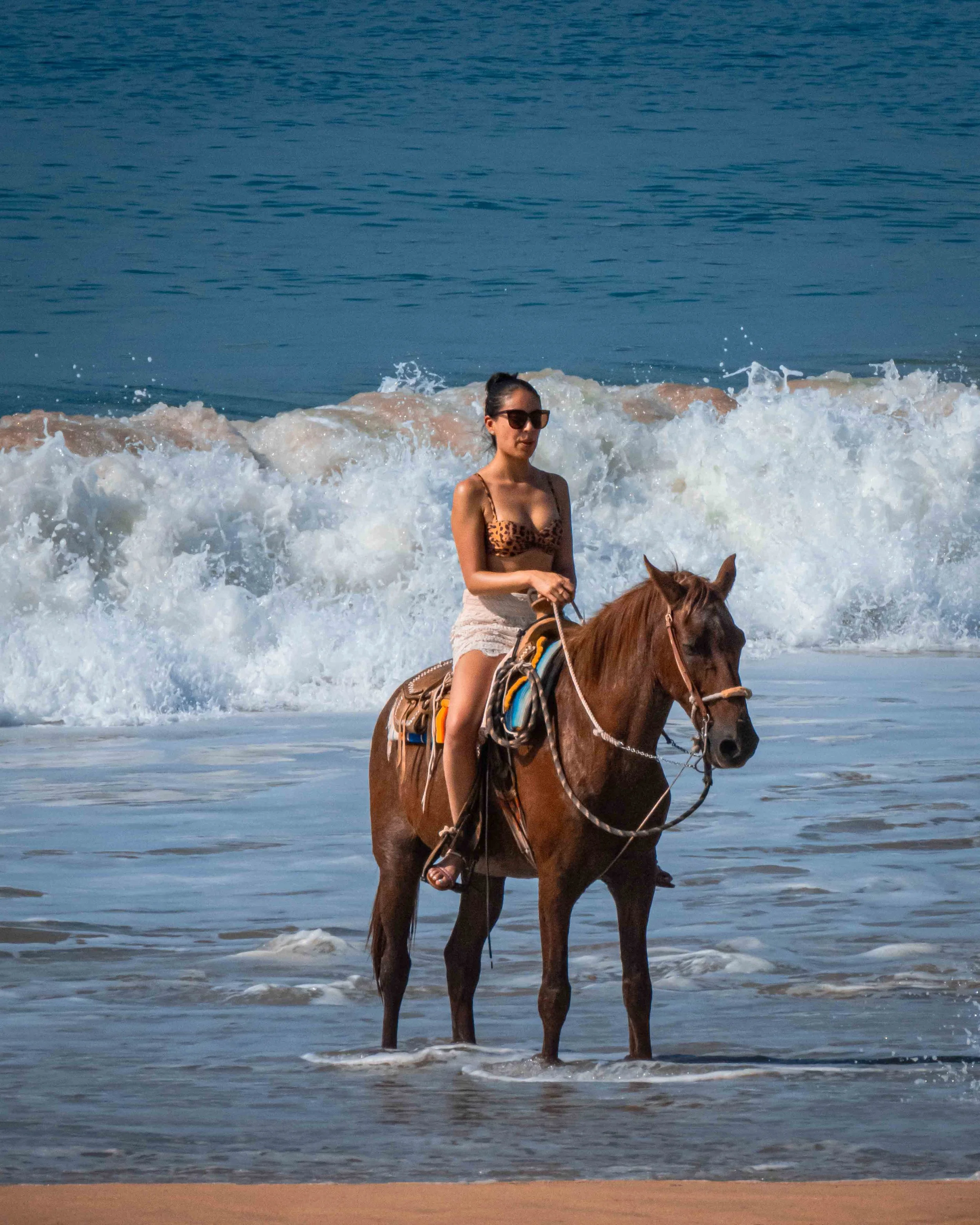 Una mujer montando un caballo en la orilla del mar, con olas rompiendo en el fondo, usando gafas de sol y ropa de verano. hotel baxar pie de la cuesta