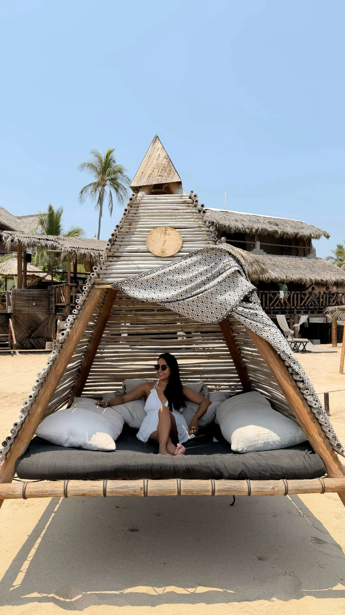 Mujer con vestido blanco y gafas de sol relajada en una estructura de madera y cortinas en la playa. Hotel boutique playa pie de la cuesta, baXar Acapulco